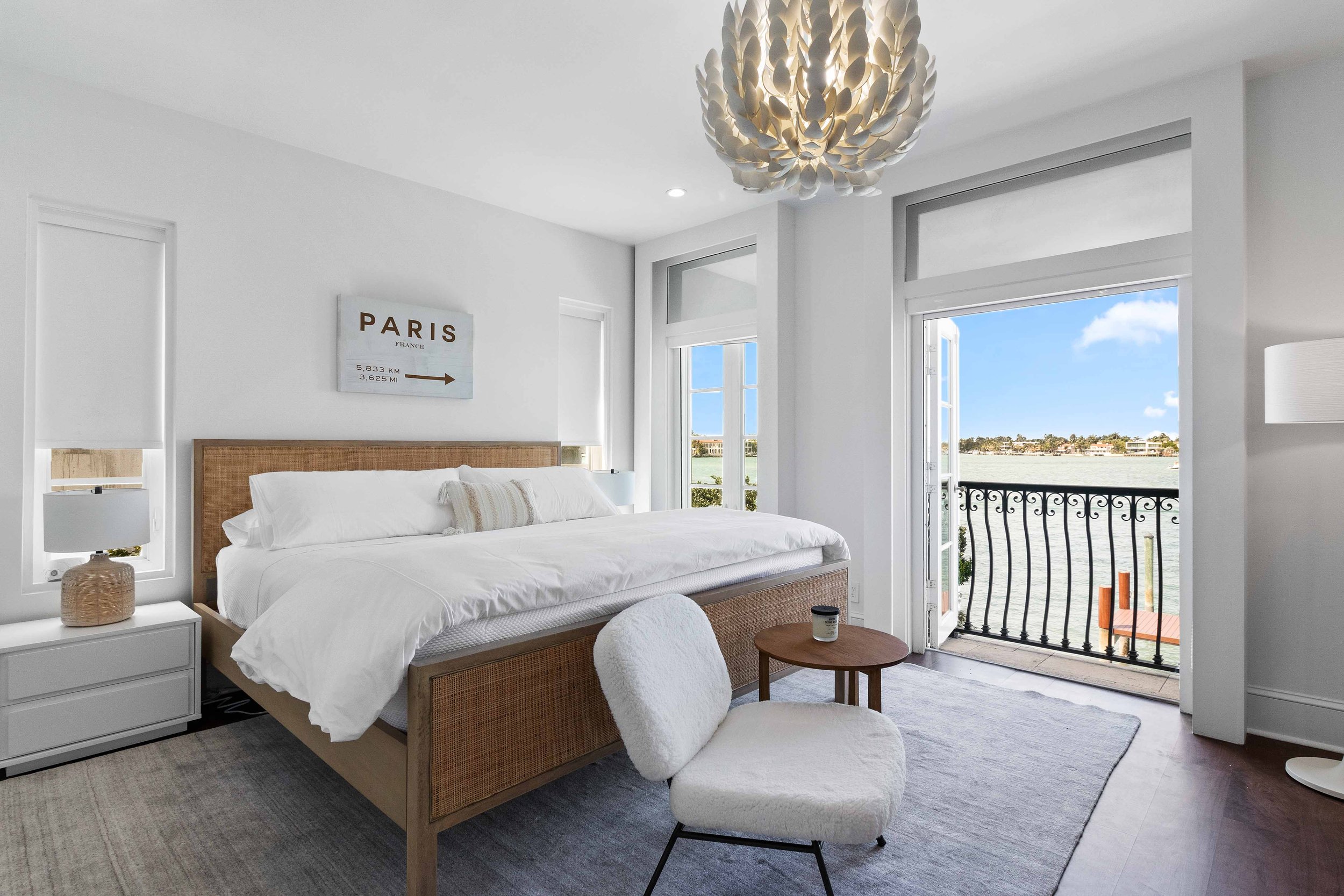 Waterfront bedroom with cane bed frame, sculptural shell chandelier, and balcony bay views on Hibiscus Island in Miami Beach, Interior Design by CURATED.