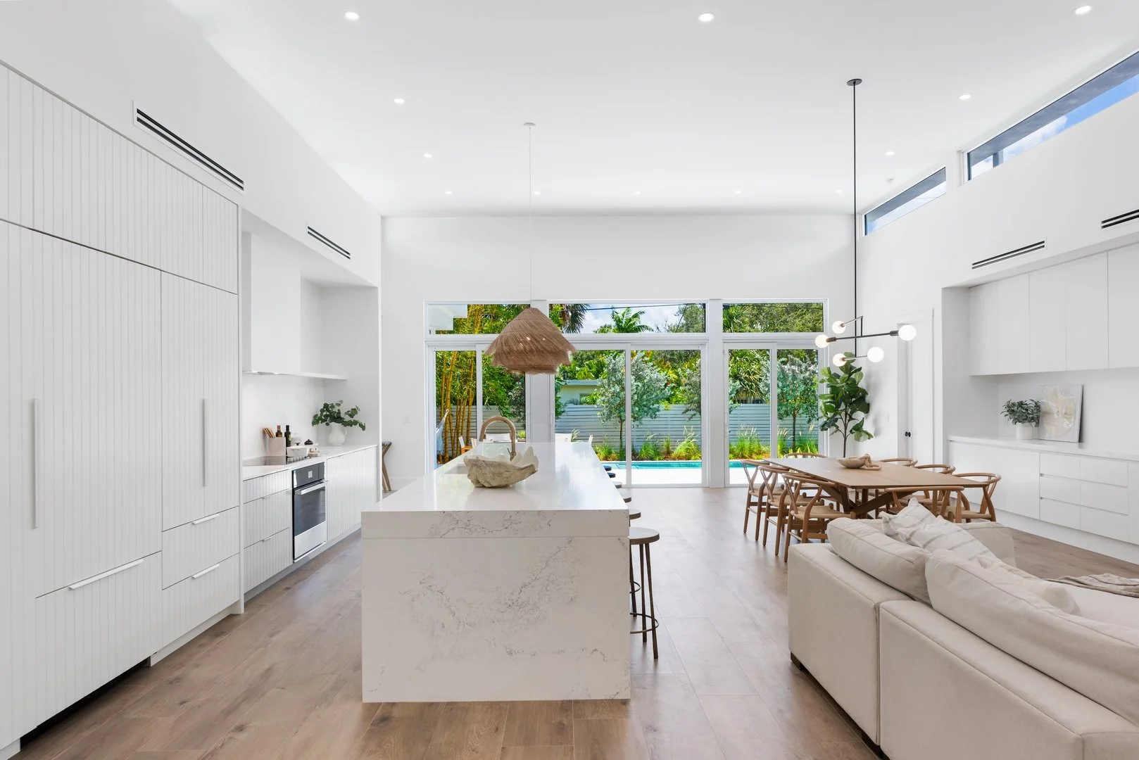 Bright open-concept kitchen and dining area with white marble waterfall island, woven pendant, and wishbone chairs in Wilton Manors, Interior Design by CURATED.