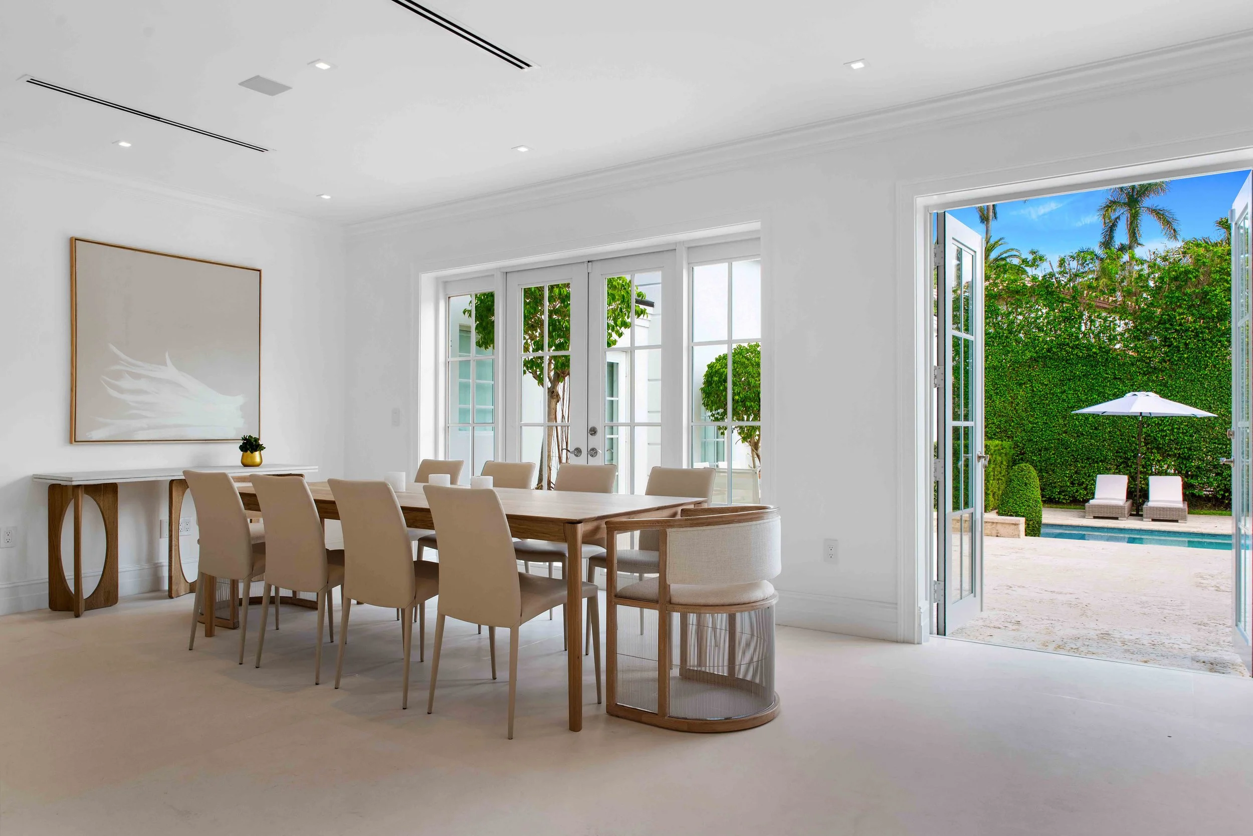 Bright dining room with light wood table, upholstered chairs, and French doors opening to the pool on La Gorce Island in Miami Beach, Interior Design by CURATED.