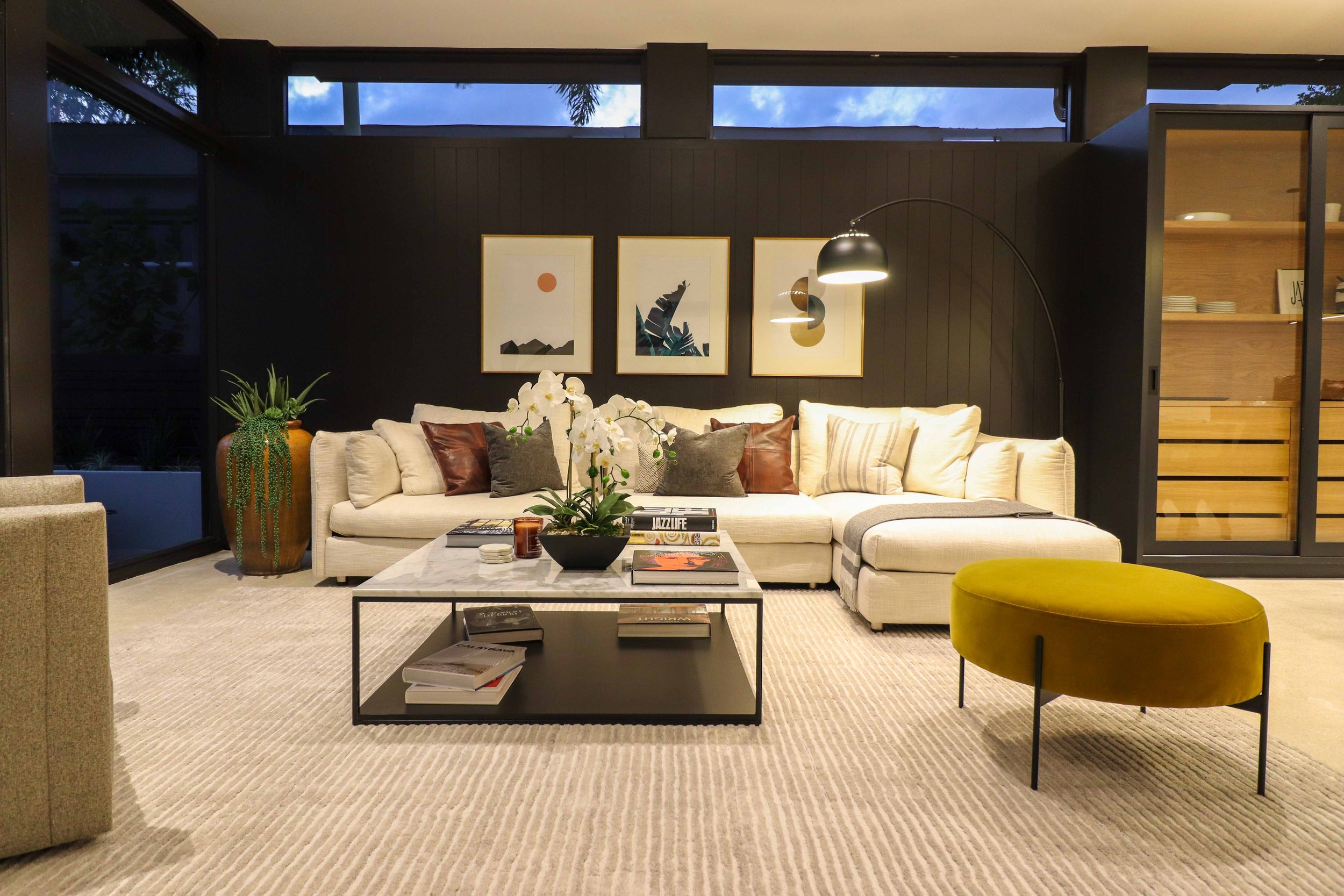 Mid-century modern living room with cream sectional, mustard velvet ottoman, black arc floor lamp, and dark paneled accent wall in Wilton Manors, Interior Design by CURATED.