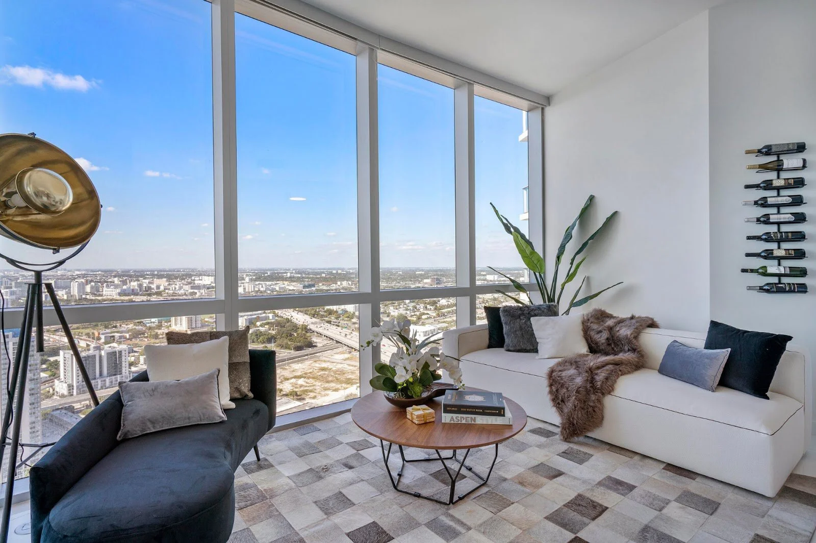 High-rise living room with navy velvet chaise, white sofa with faux fur throw, patchwork hide rug, and wall-mounted wine rack overlooking the Downtown Miami skyline, Interior Design by CURATED.