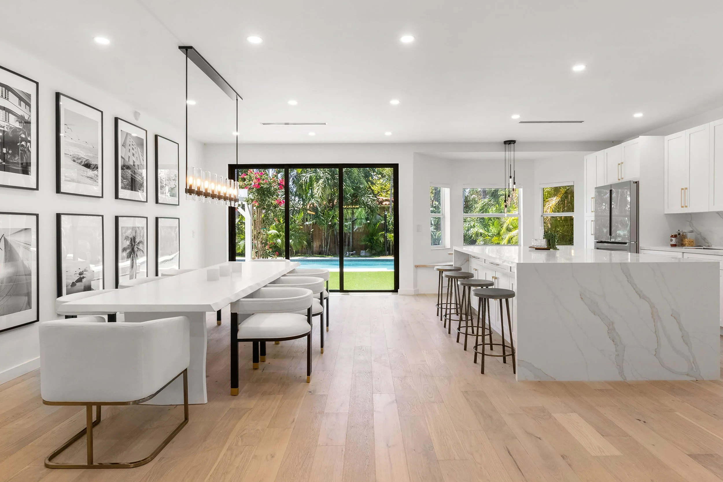 Open kitchen and dining area with long white dining table, black-and-white photo gallery wall, and marble waterfall island in Miami Beach, Interior Design by CURATED.