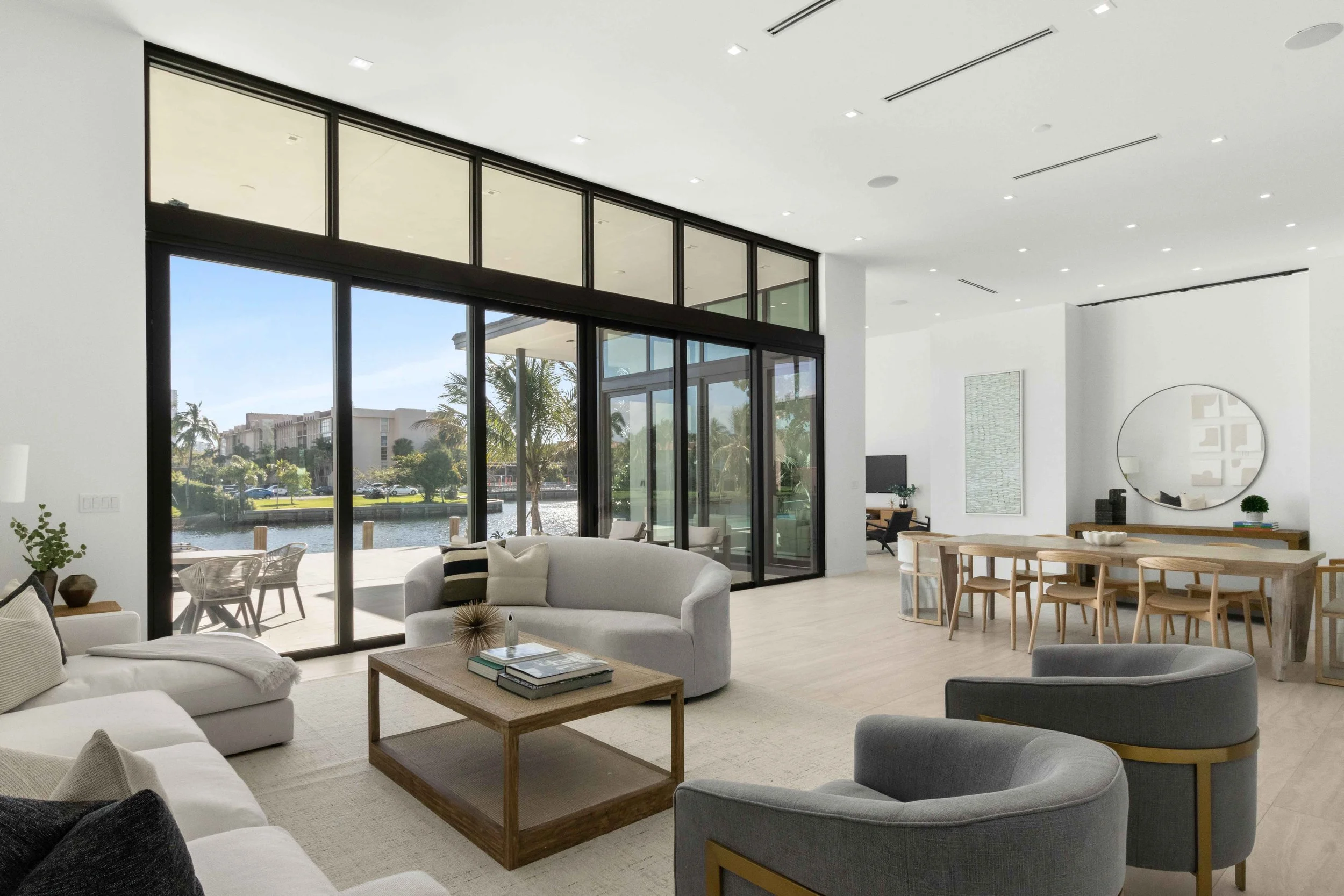 Double-height waterfront living room with curved white sofa, gray barrel swivel chairs, and floor-to-ceiling glass on Diplomat Parkway, Interior Design by CURATED.