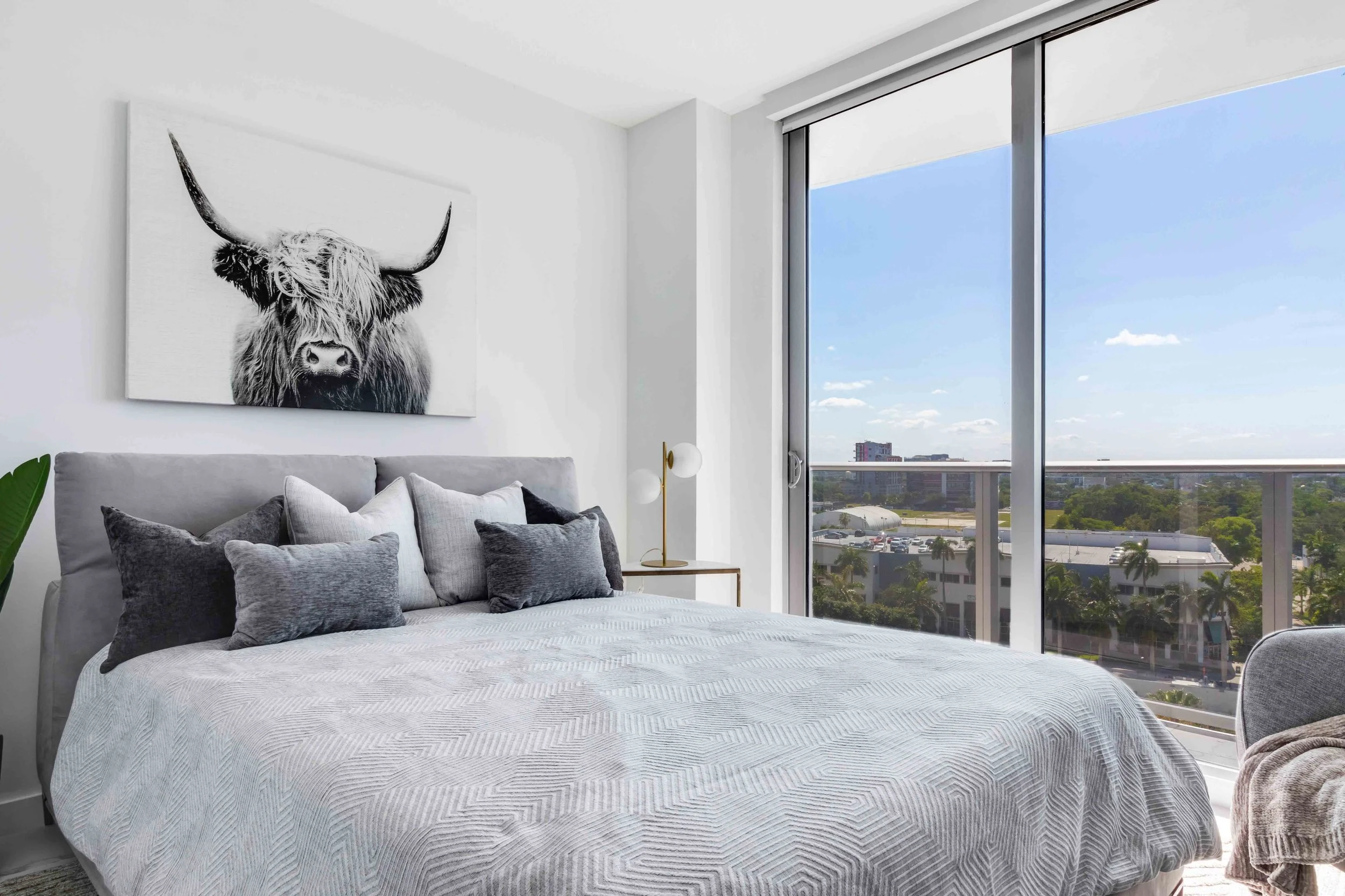 Contemporary bedroom with gray upholstered bed, layered charcoal pillows, black-and-white Highland cow artwork, and floor-to-ceiling city views in Downtown Miami, Interior Design by CURATED.