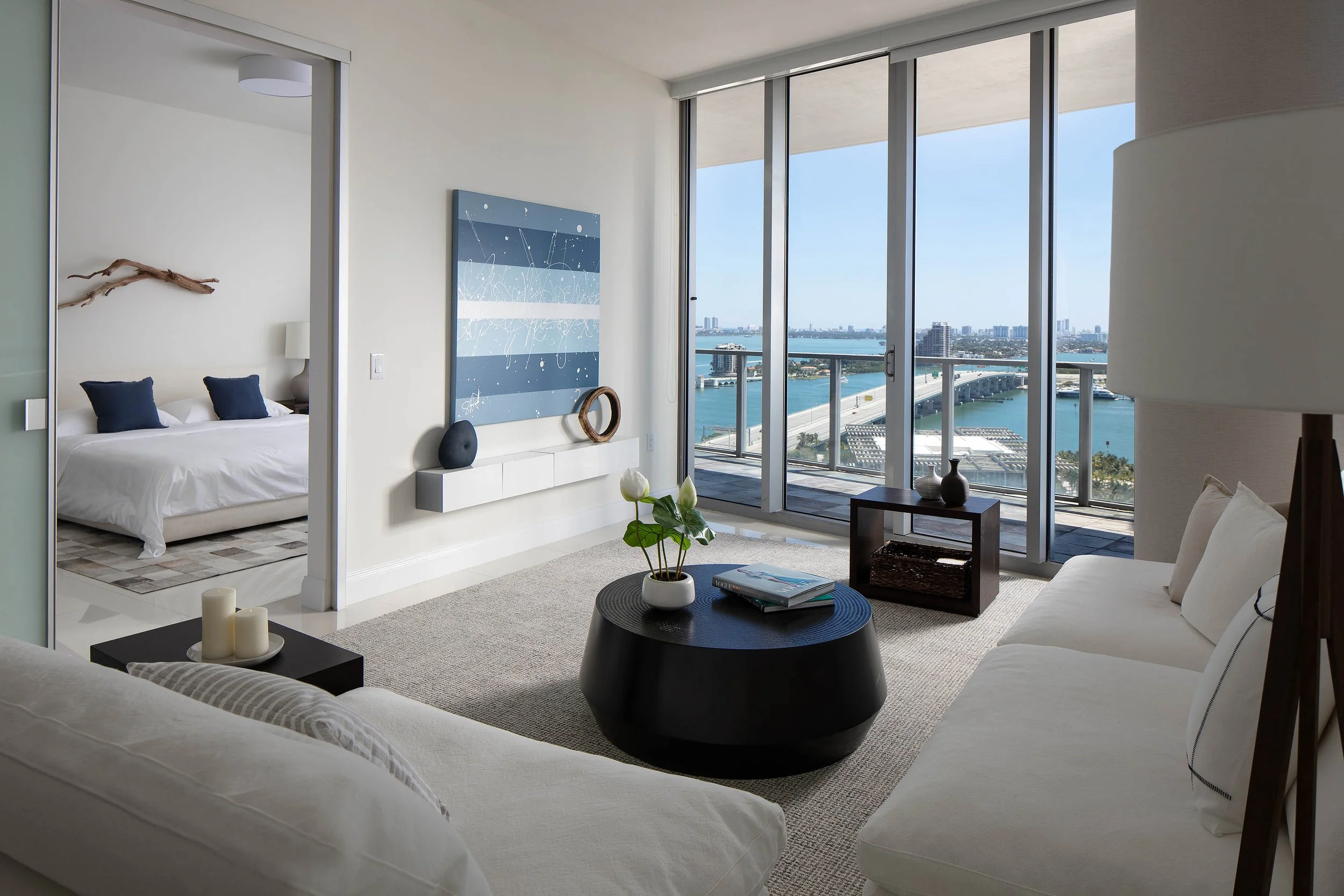 Minimalist living room with white sofas, round black drum coffee table, blue striped artwork, and floor-to-ceiling bay views in South Florida, Interior Design by CURATED.