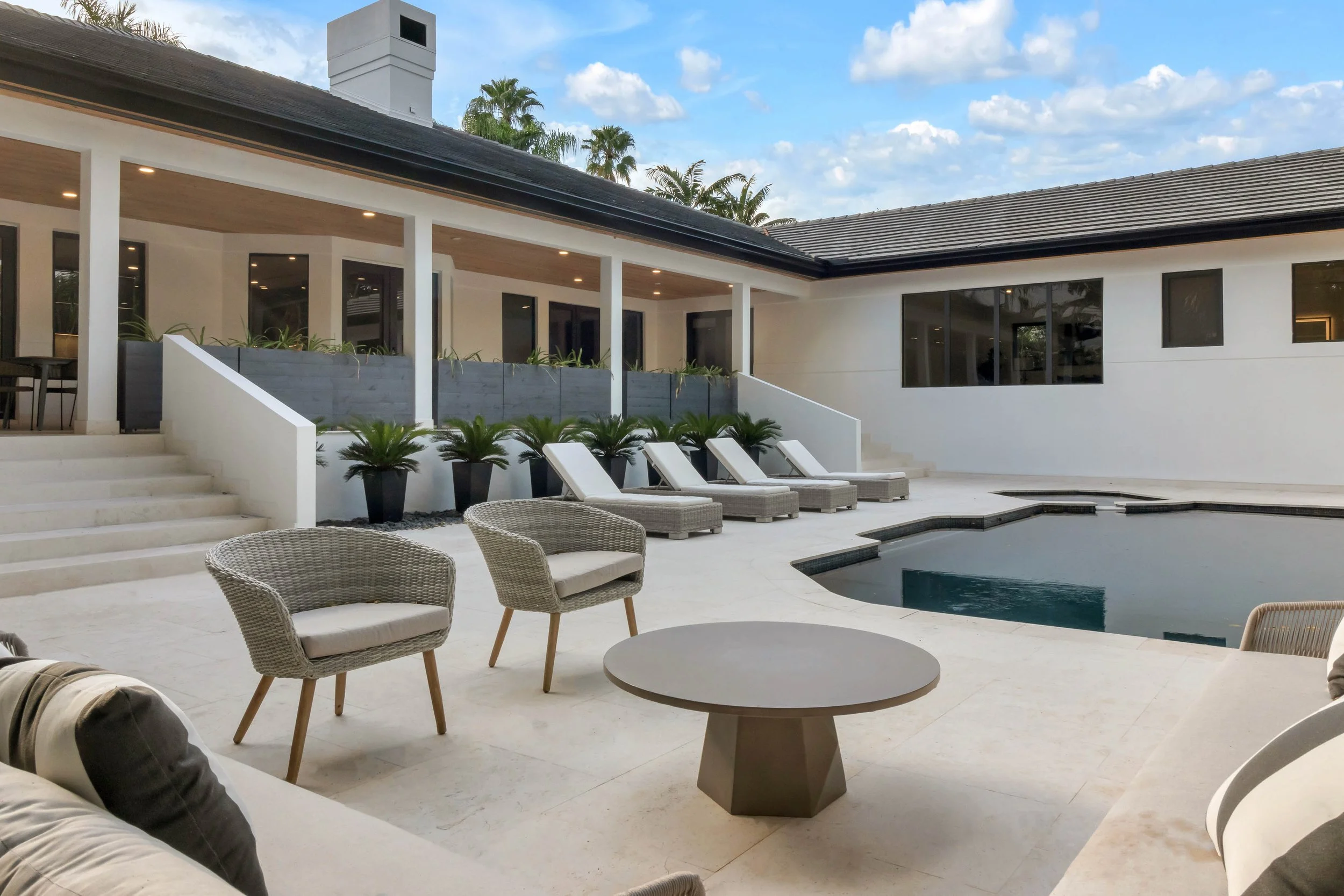 Pool deck with woven wicker lounge chairs, round concrete coffee table, and row of chaise loungers at Cocoplum in Coral Gables, Interior Design by CURATED.