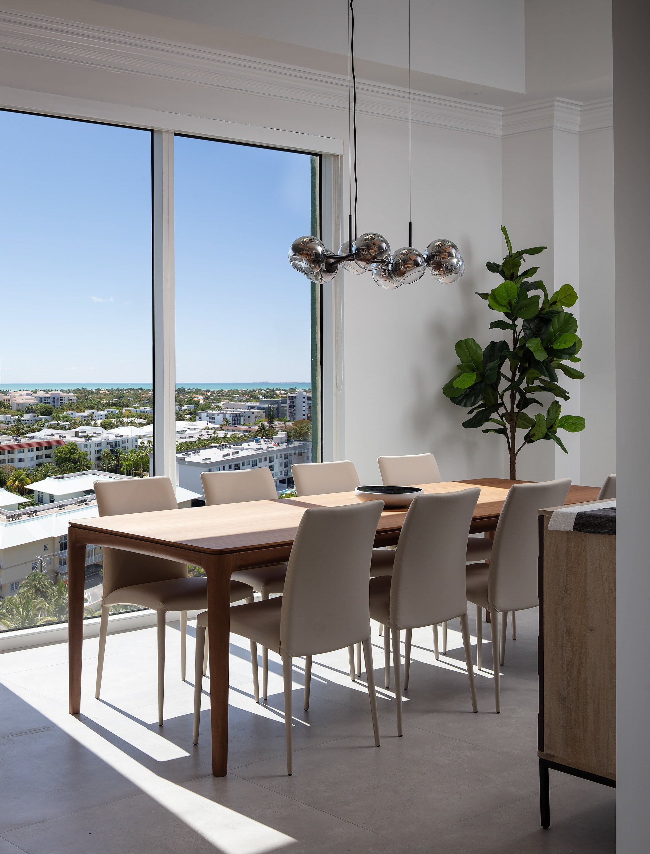 Penthouse dining room with walnut table, beige upholstered chairs, smoked glass globe chandelier, and floor-to-ceiling ocean views in Key Biscayne, Interior Design by CURATED.
