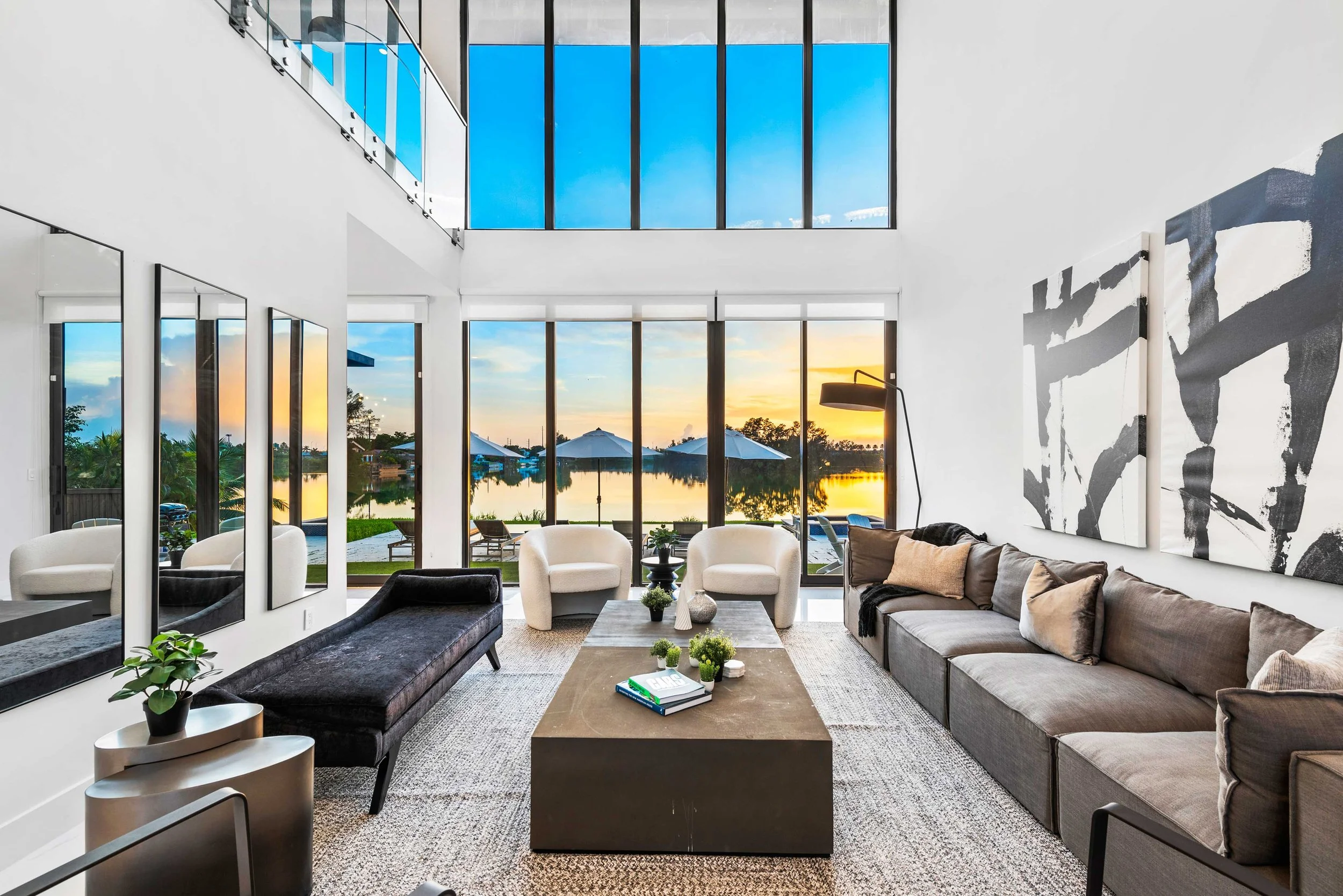 Double-height living room with gray sectional, black chaise, bold black-and-white abstract art, and floor-to-ceiling lake views in South Miami, Interior Design by CURATED.