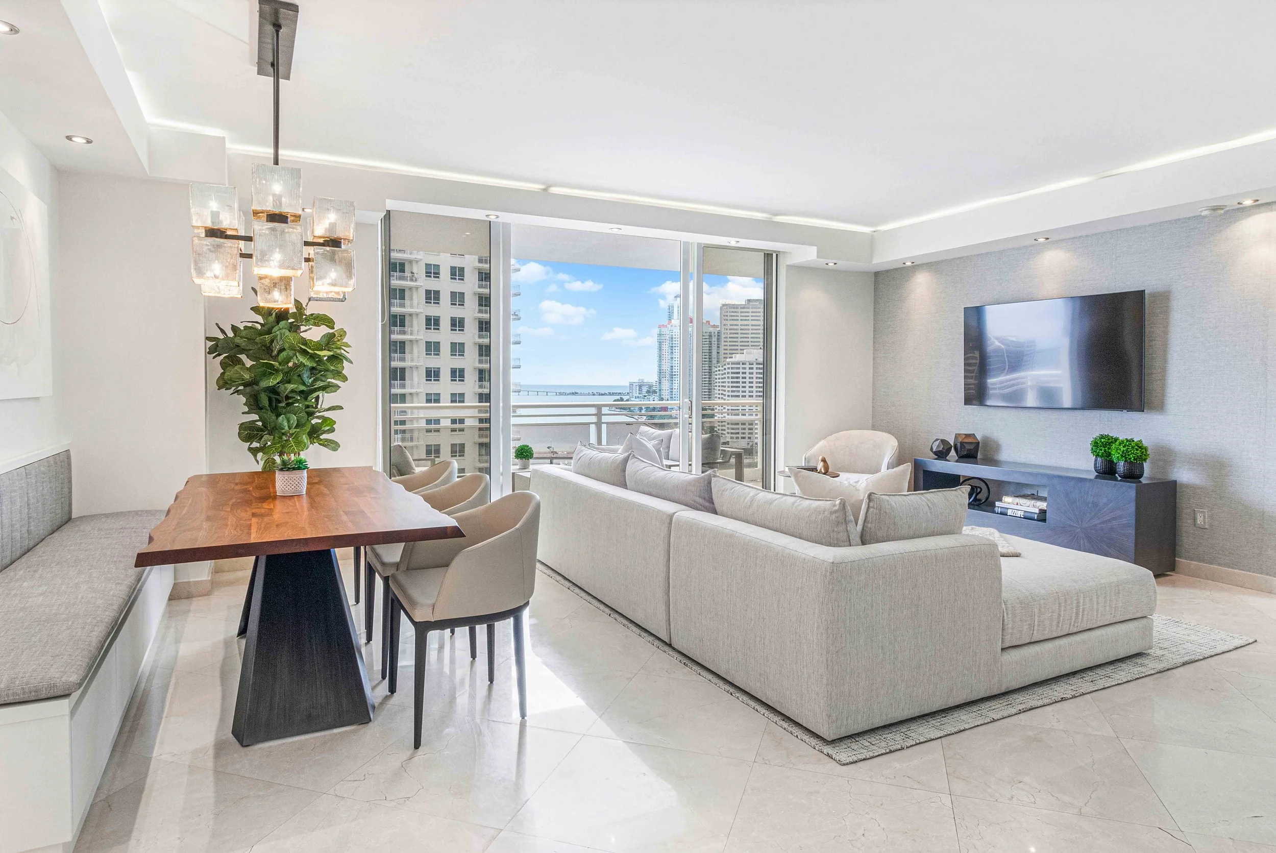Open living and dining area with gray sectional, live-edge wood dining table, built-in banquette, and glass cube chandelier at Carbonell on Brickell Key in Miami, Interior Design by CURATED.
