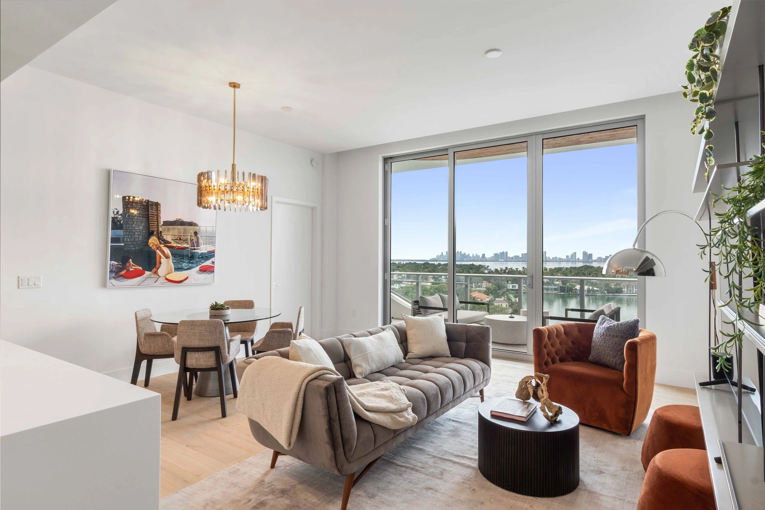 Bayfront living room with tufted gray sofa, rust velvet armchair, and brass crystal chandelier over the dining area at Monaco Yacht Club & Residences in Miami Beach, Interior Design by CURATED.