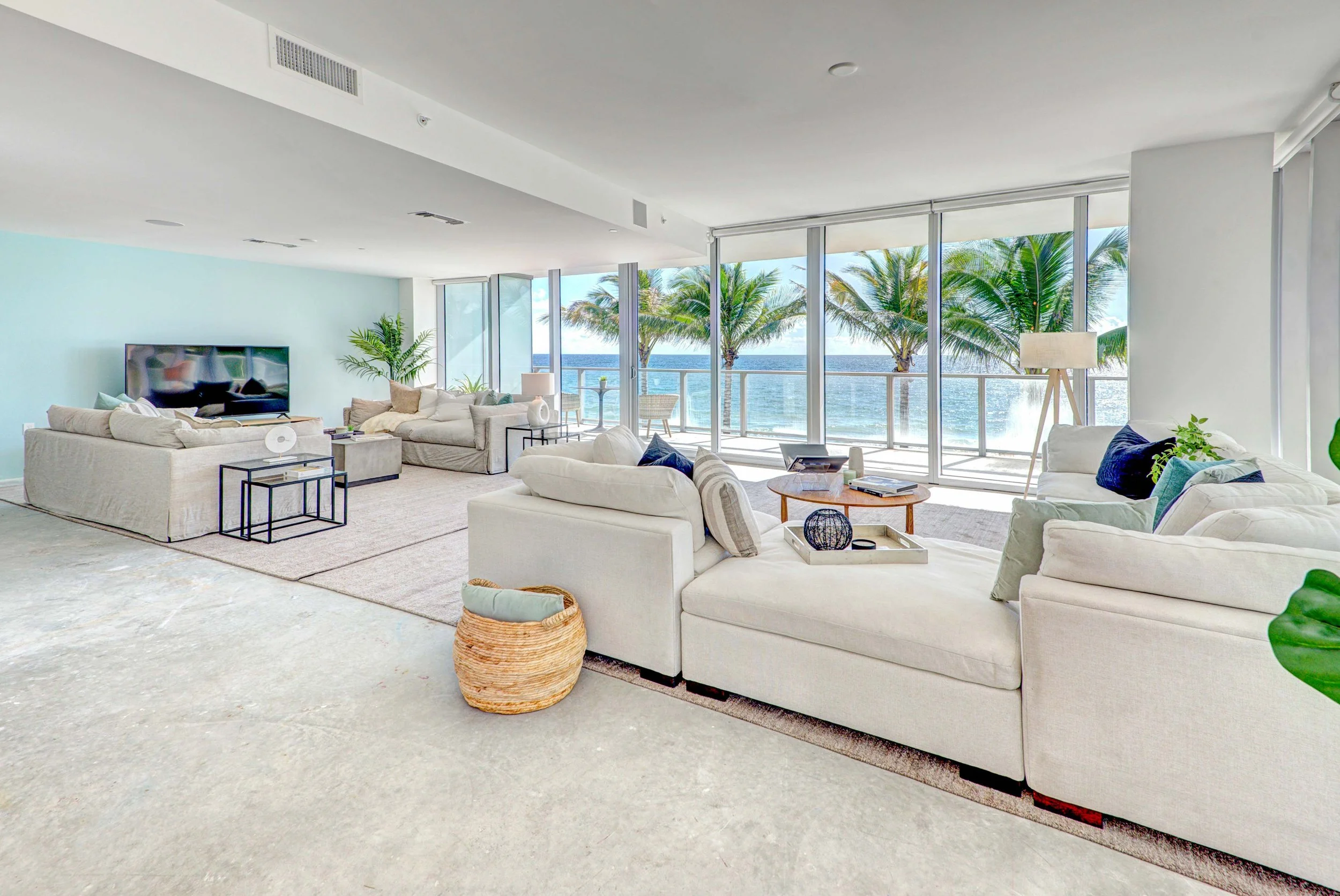 Oceanfront living room with white slipcovered sectional, navy and seafoam accent pillows, woven basket, and floor-to-ceiling palm and ocean views in Fort Lauderdale Beach, Interior Design by CURATED.