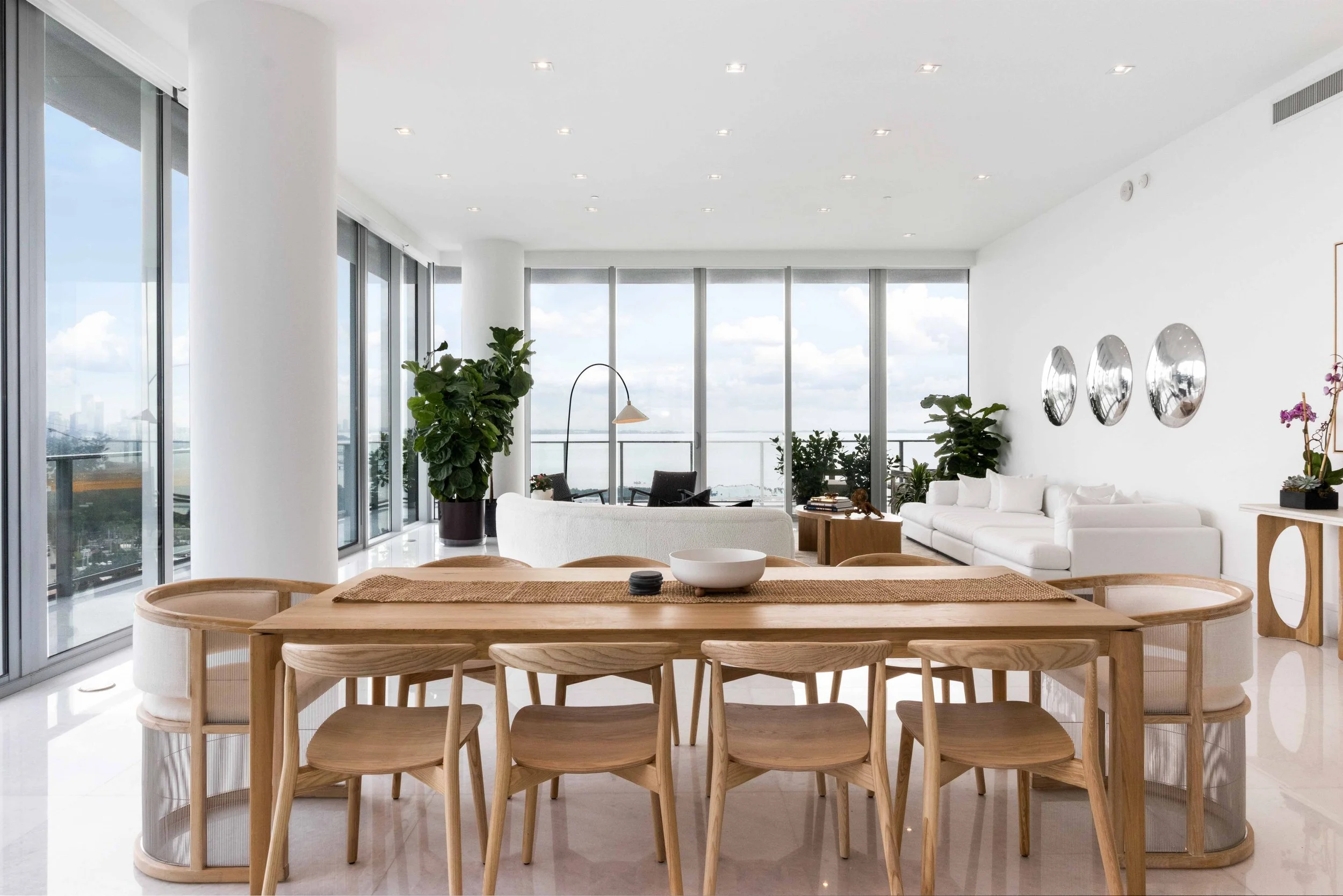 Penthouse dining area with light oak table and chairs, sculptural mirror trio, and floor-to-ceiling bay views in Coconut Grove, Interior Design by CURATED.
