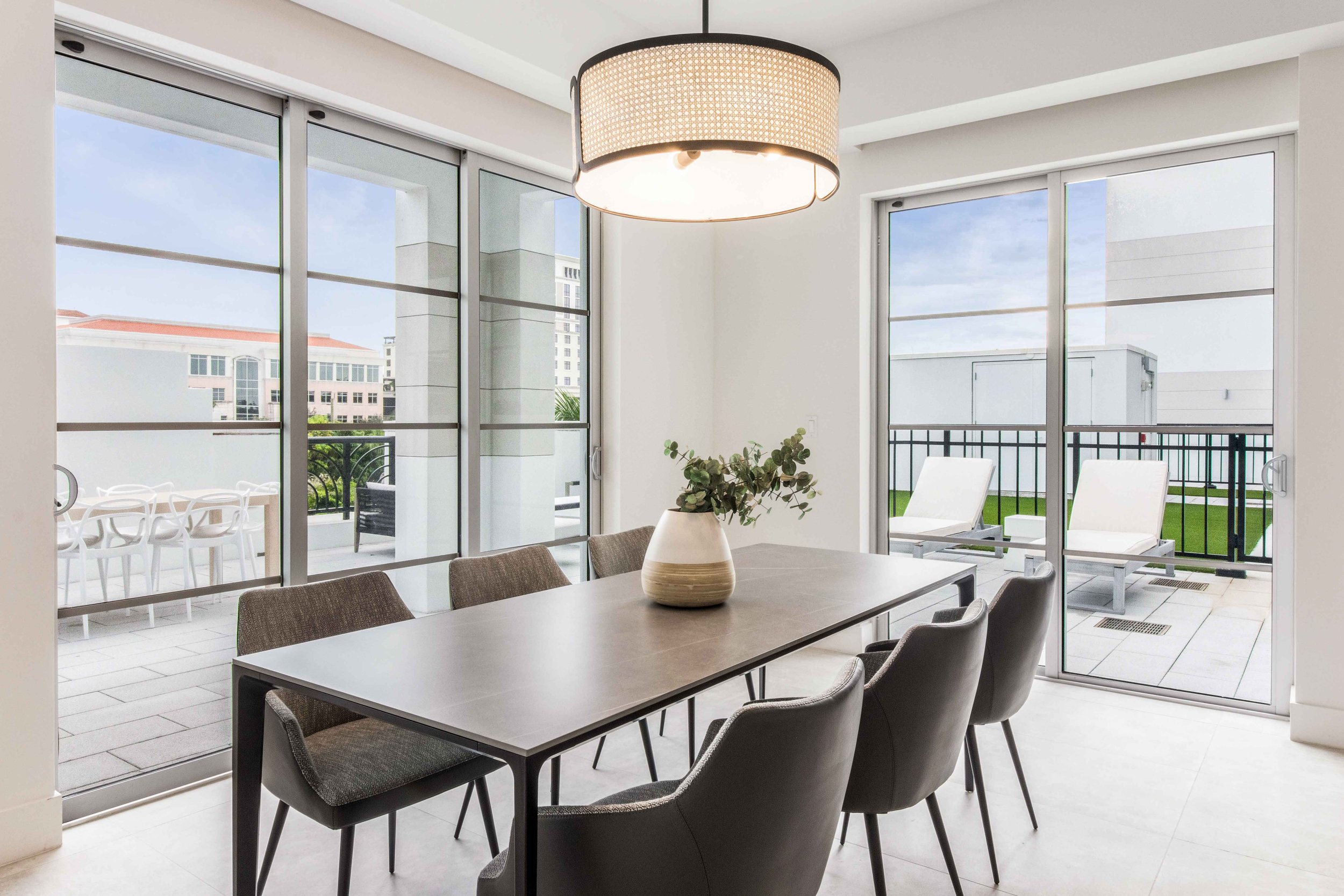 Contemporary dining room with dark rectangular table, charcoal upholstered chairs, and woven cane drum pendant opening to the terrace in Boca Raton, Interior Design by CURATED.