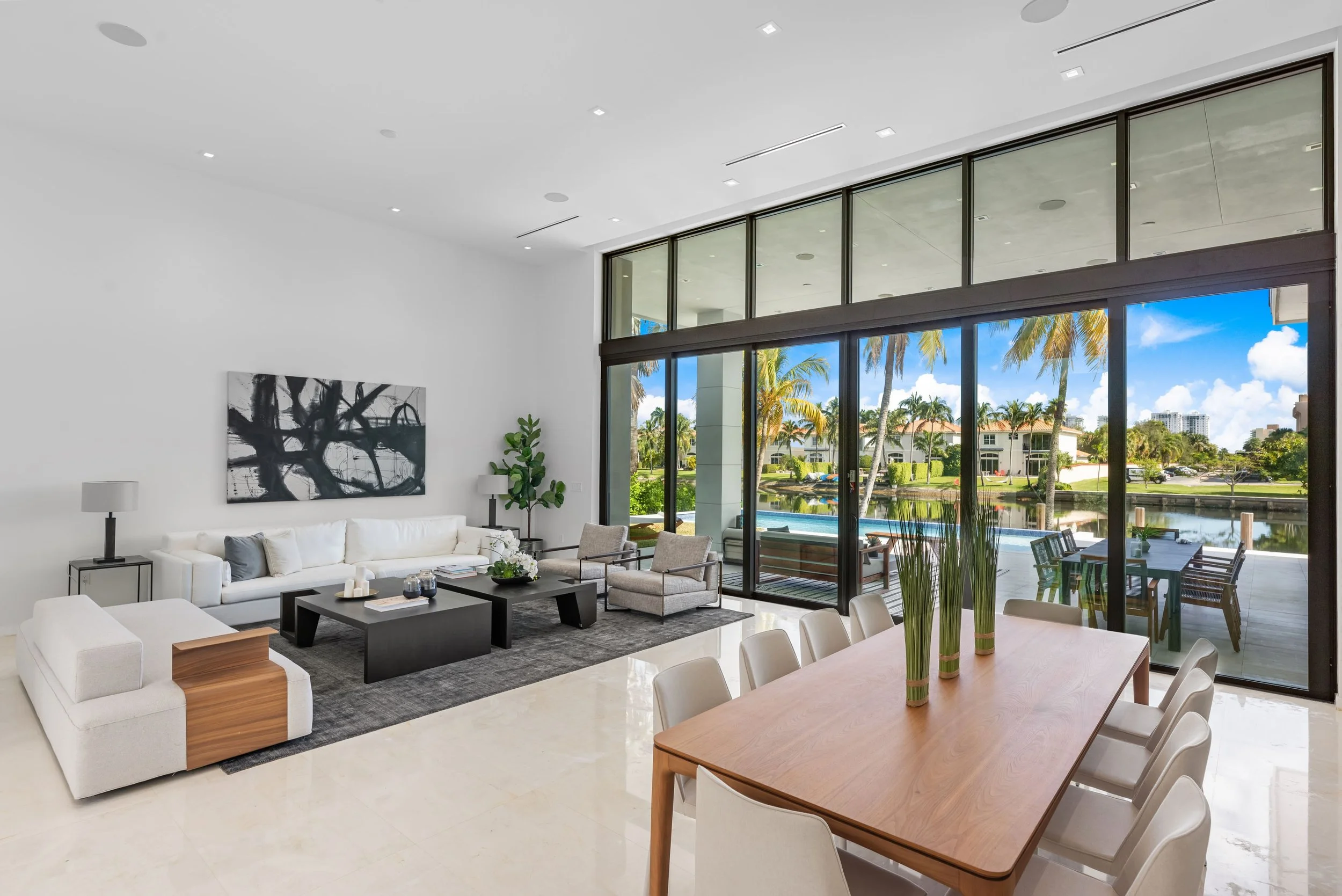 Open living and dining area with white sofas, walnut dining table, dark coffee tables, and floor-to-ceiling glass overlooking the pool and waterway in Hallandale Beach, Interior Design by CURATED.