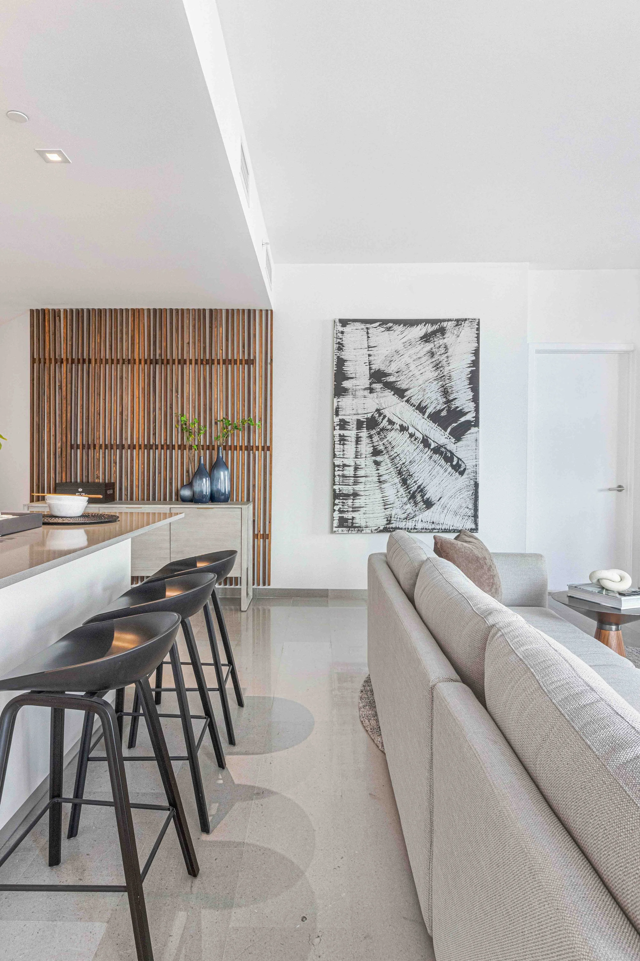 Contemporary living and kitchen area with black counter stools, slatted wood wall panel, and black-and-white abstract artwork at Reach Brickell City Centre in Miami, Interior Design by CURATED.