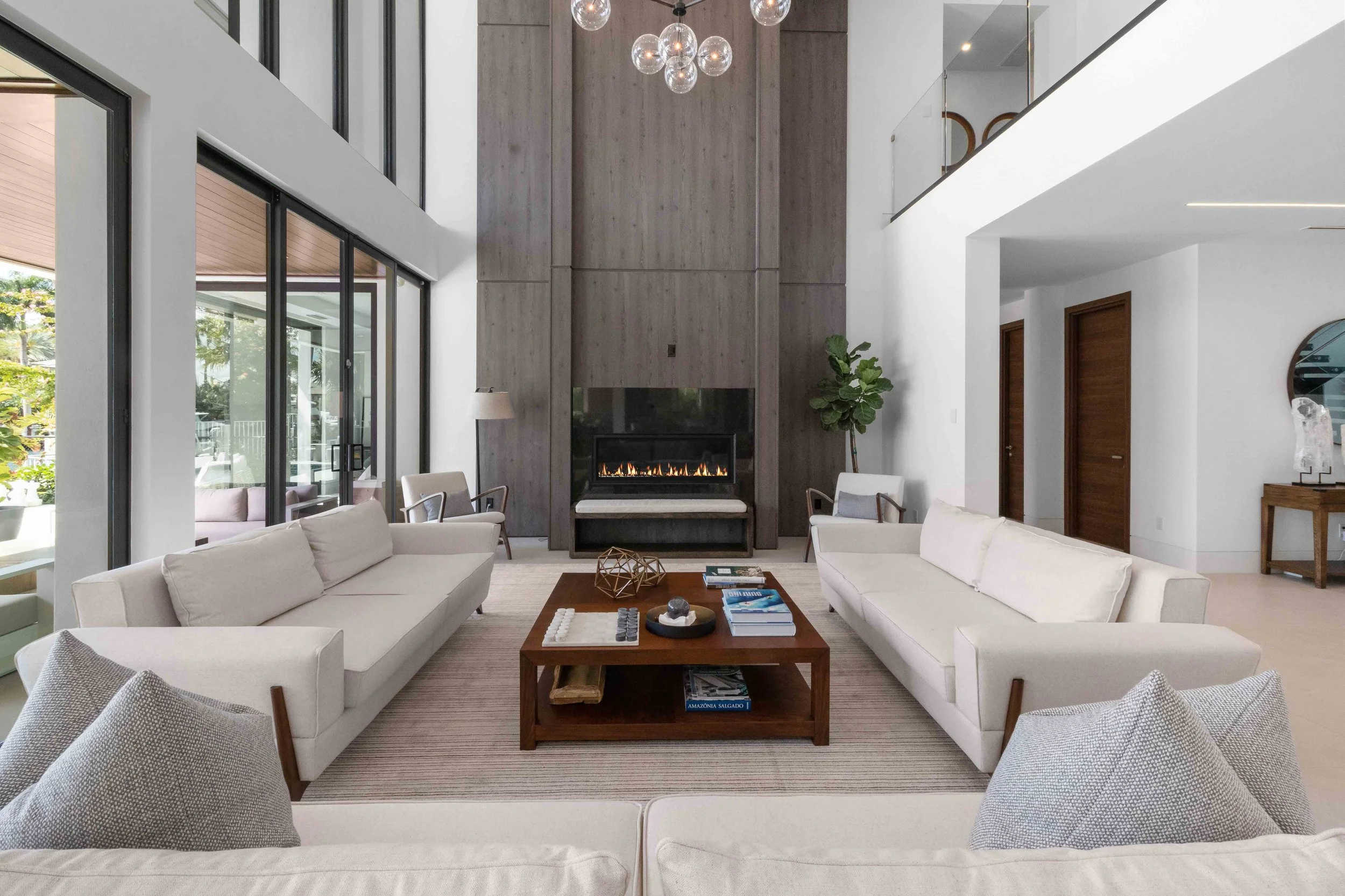 Double-height living room with facing white sofas, wood-paneled fireplace wall, and glass bubble chandelier at Bay Harbor in Fort Lauderdale, Interior Design by CURATED.