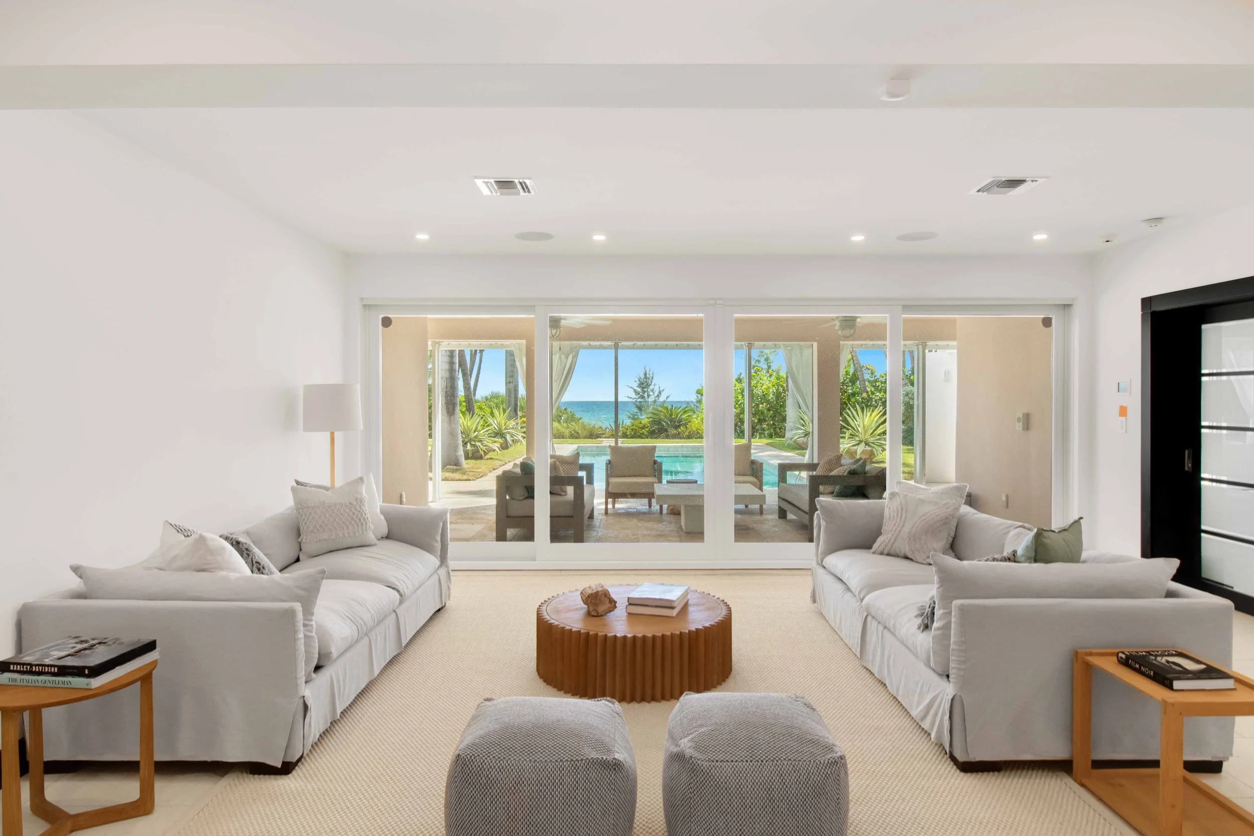 Beachfront living room with facing slipcovered sofas, round fluted wood coffee table, and woven poufs overlooking the pool and ocean in Golden Beach, Interior Design by CURATED.