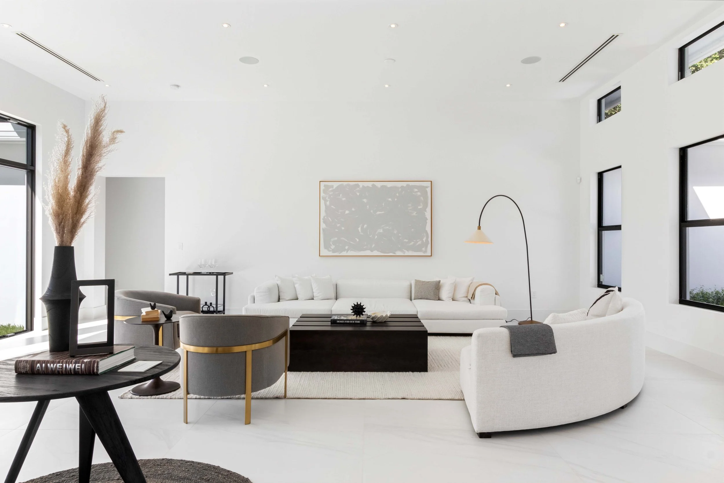 Minimalist living room with curved white sofa, gray and brass lounge chairs, and dark wood coffee table in Coral Gables, Interior Design by CURATED.