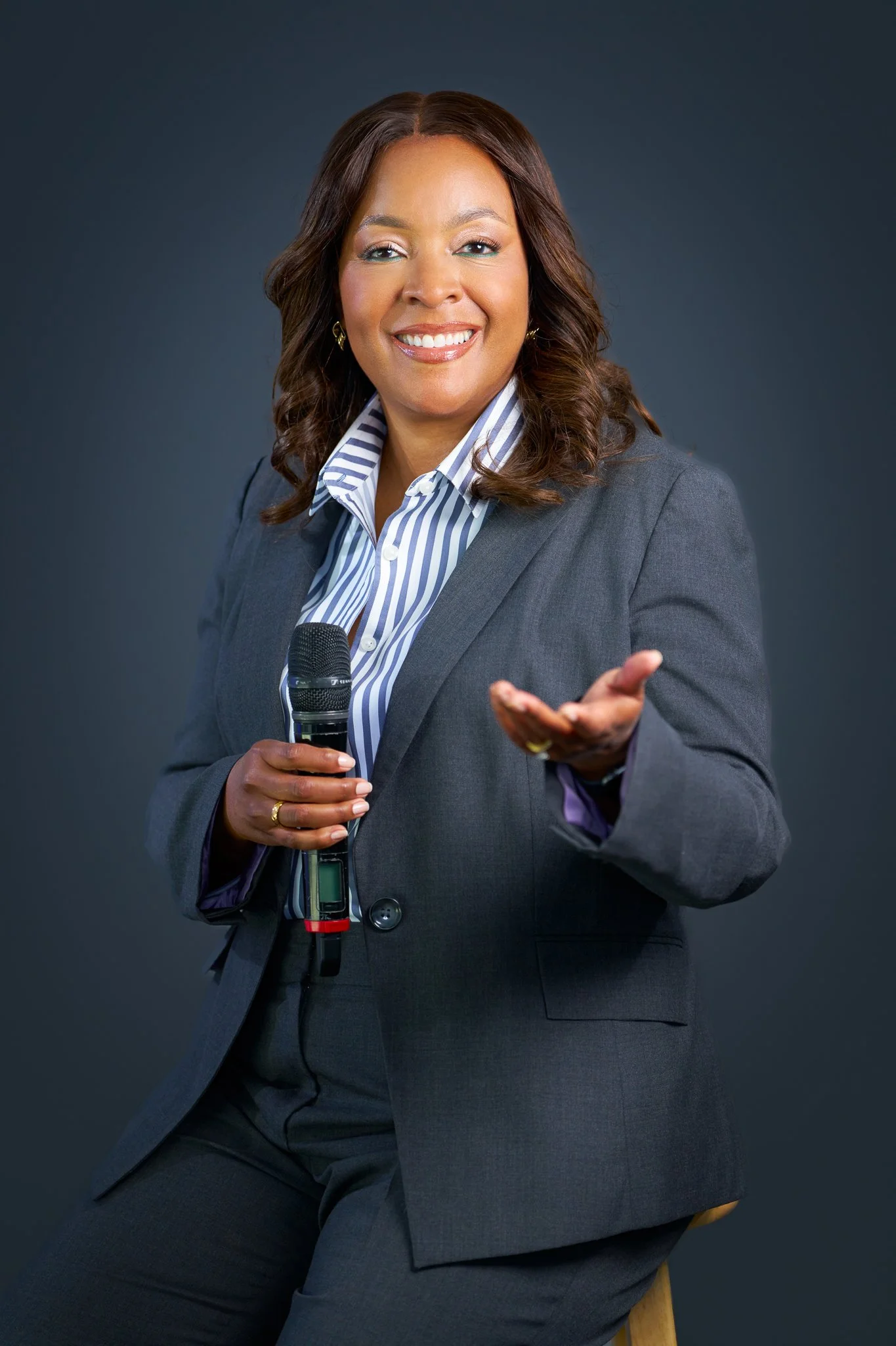A woman in a gray business suit, holding a microphone, smiling, seated against a dark background.