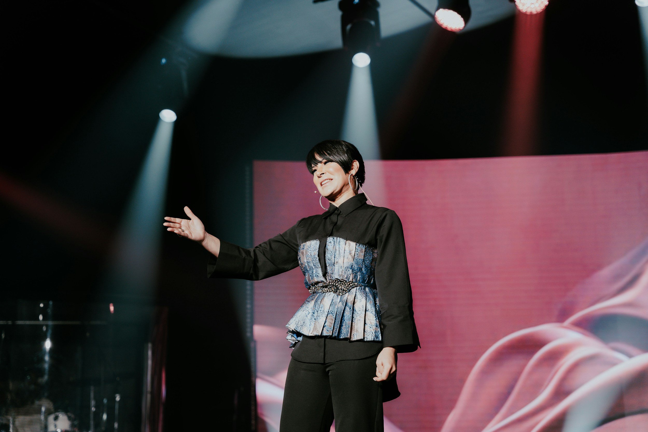 Woman with short black hair smiling on stage, wearing black and blue patterned outfit with a peplum waist, standing in front of a pink backdrop, with stage lights above.