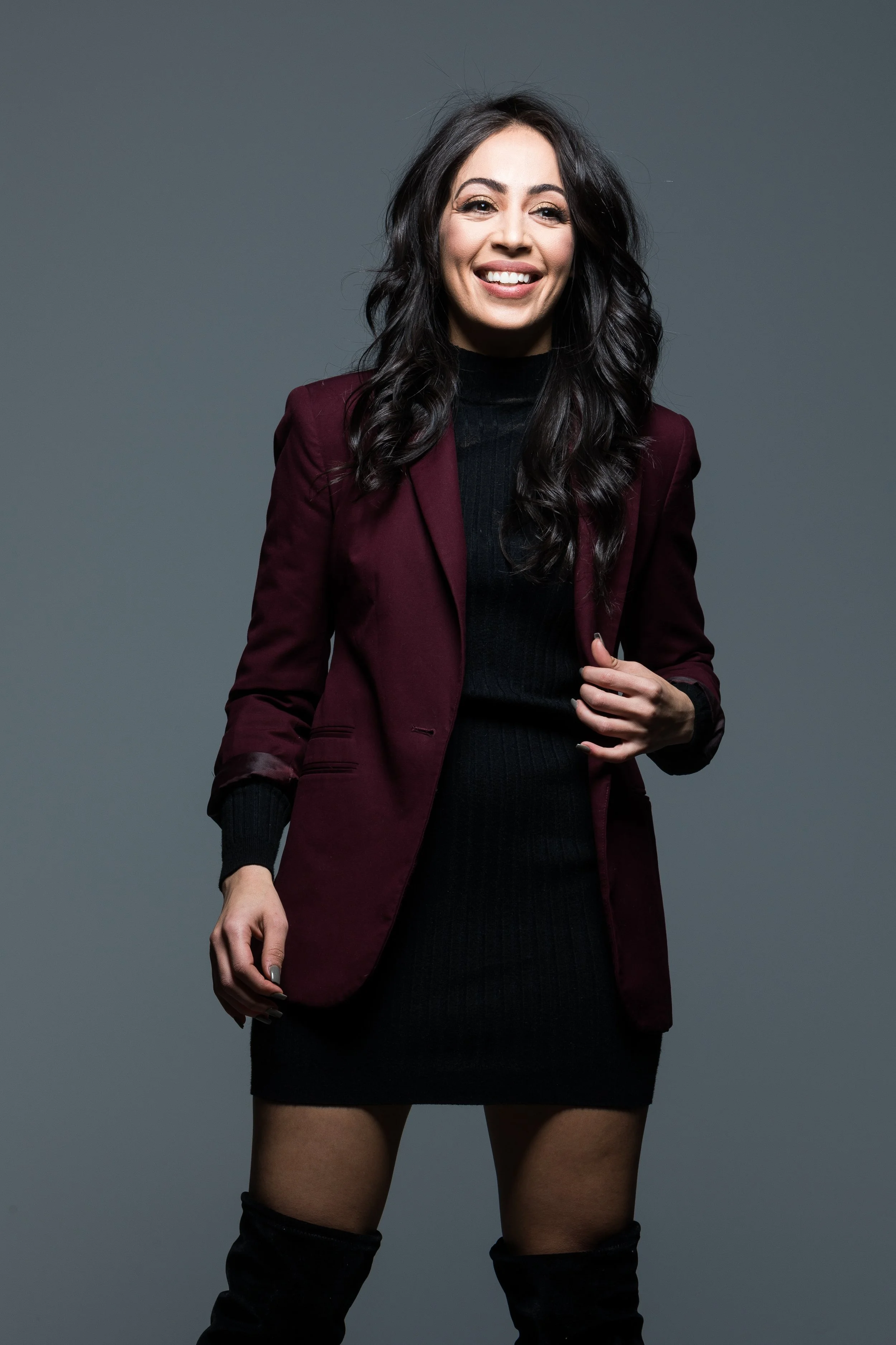 A young woman with long dark hair, smiling, wearing a black dress, black knee-high boots, and a maroon blazer, standing against a gray background.