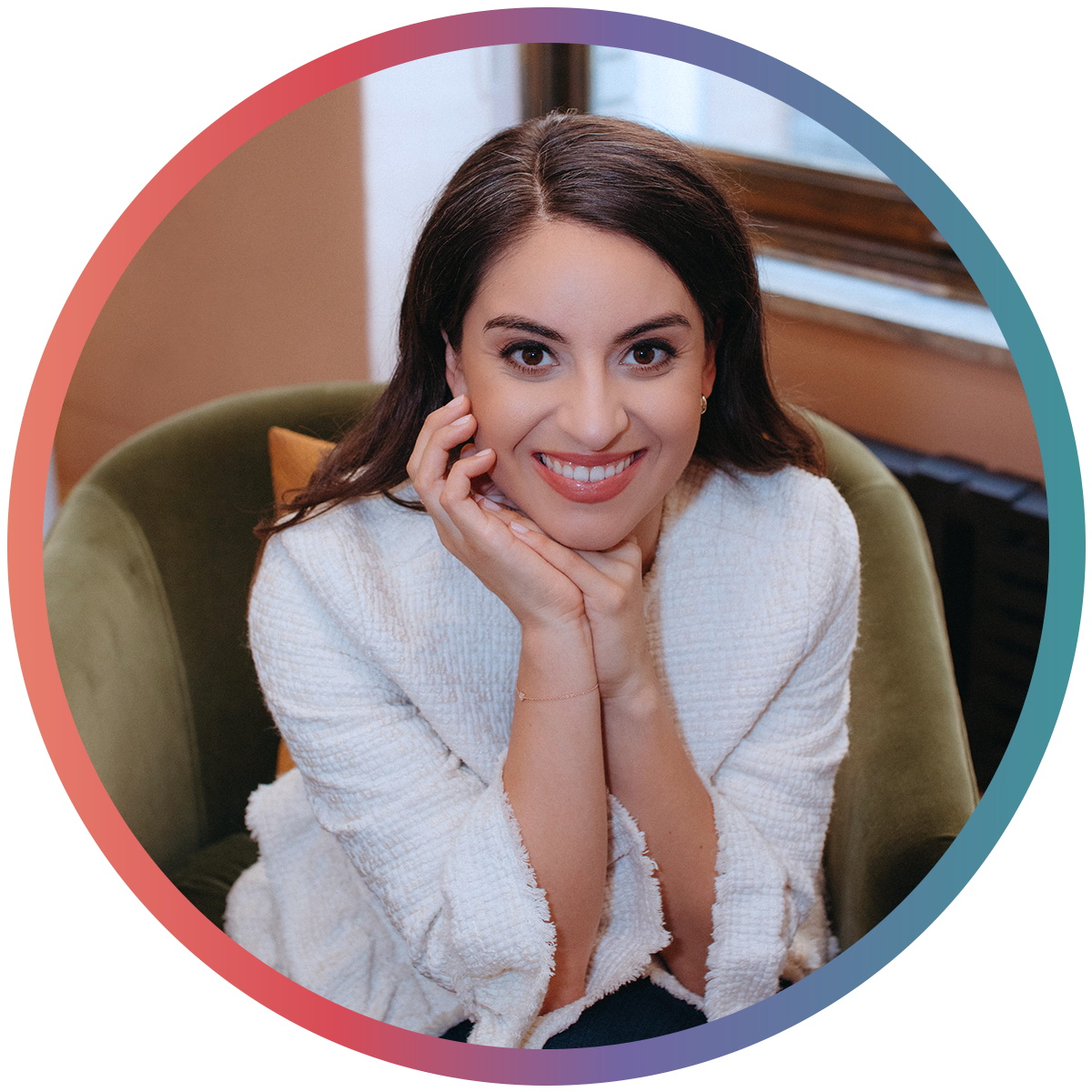 A smiling woman with dark hair wearing a white textured top, sitting on a green armchair near a window.