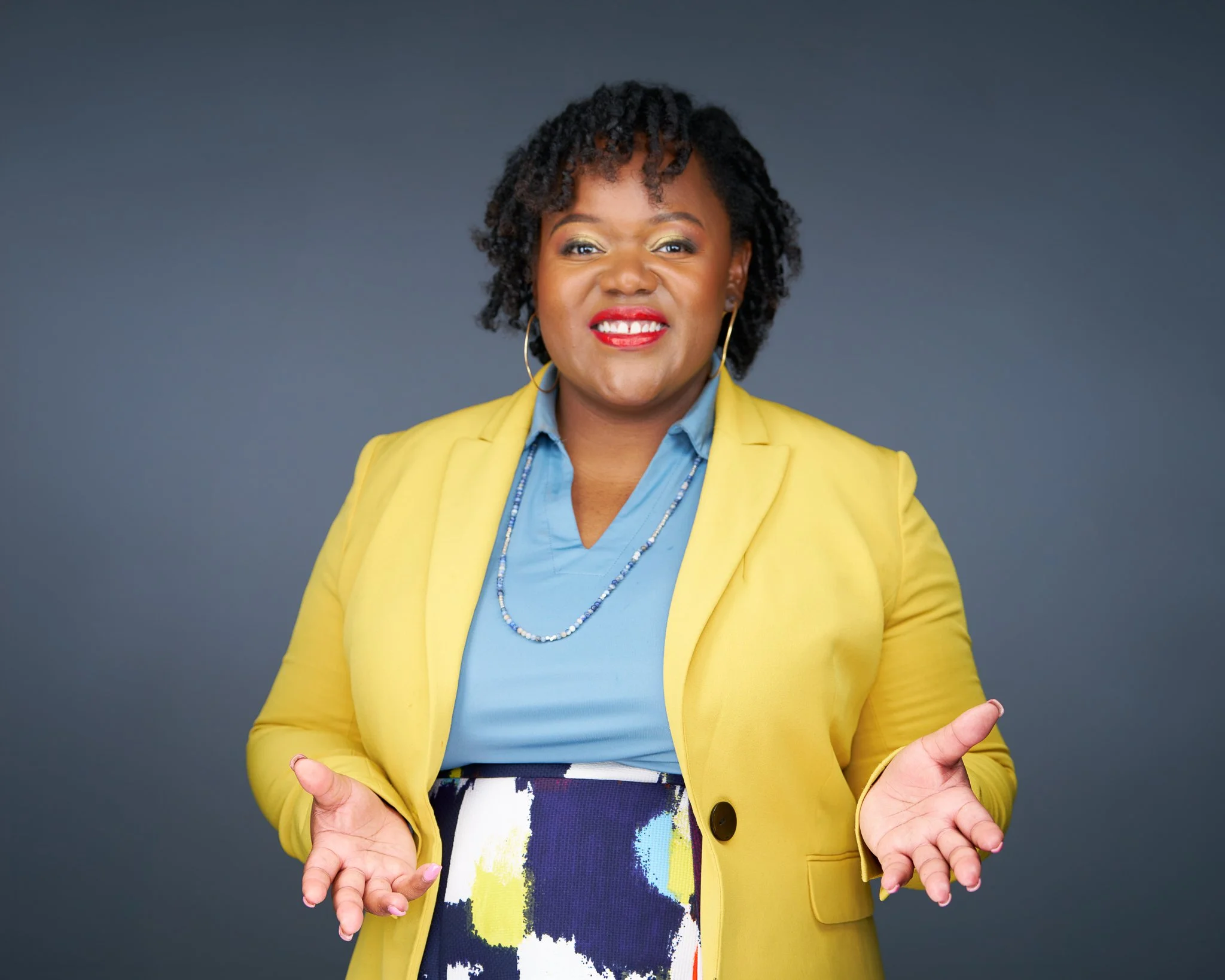A smiling African American woman with short curly hair, wearing a yellow blazer over a blue blouse, accessorized with hoop earrings and a necklace, standing against a gray background with her hands open in a welcoming gesture.