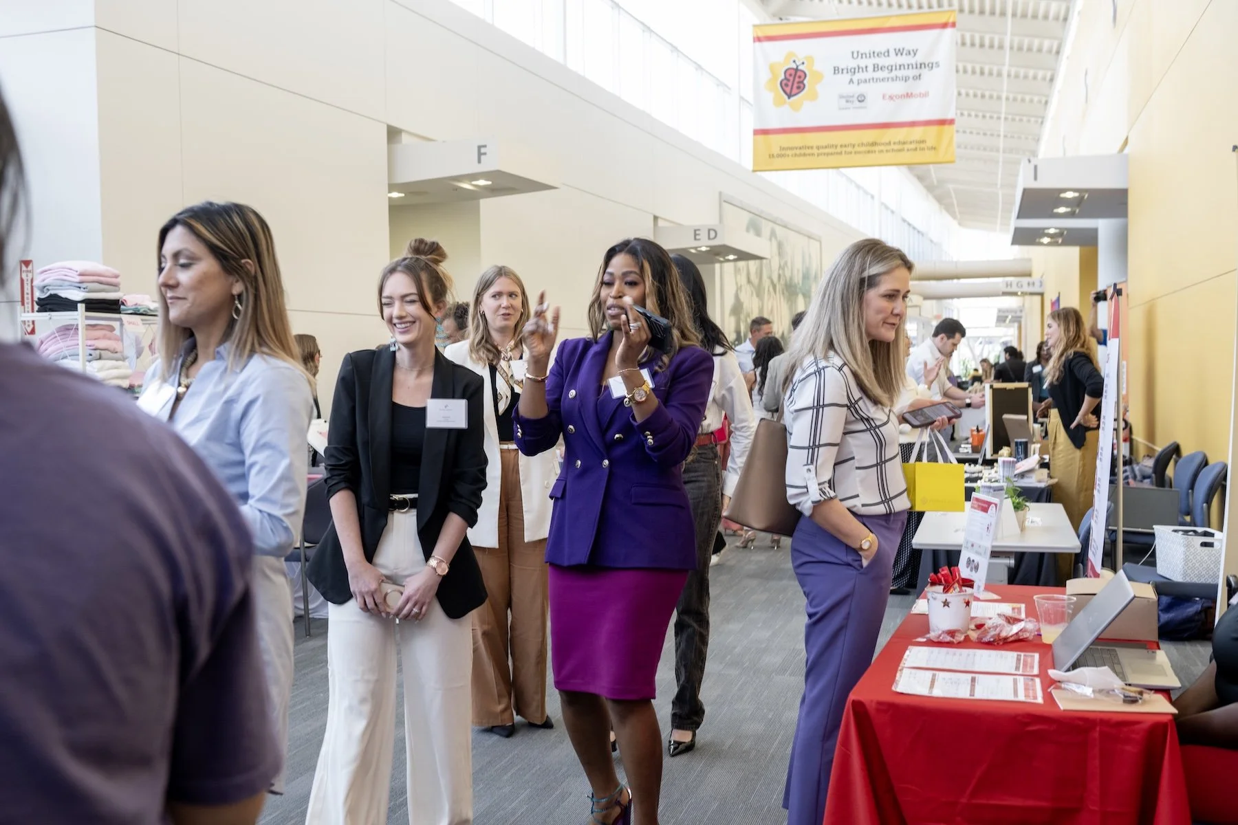 Group of professional women at an indoor networking event or conference, some smiling, others engaging with booths or booths, with a banner overhead reading 'United Way Bright Beginnings'.