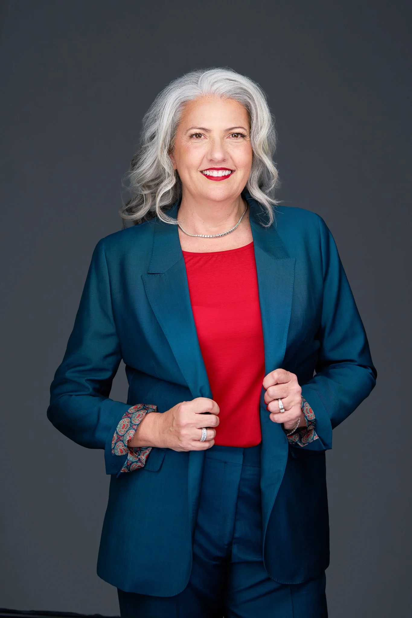 A portrait of a smiling woman with gray hair, wearing a teal business suit over a red top, standing against a gray background.