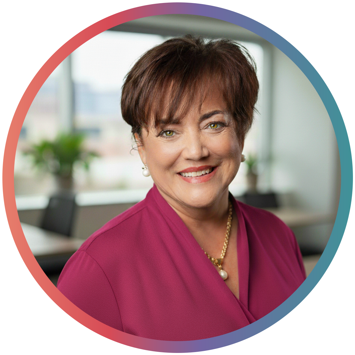 A smiling woman with short brown hair wearing a purple blouse and pearl necklace, sitting in a modern office with large windows and plants.