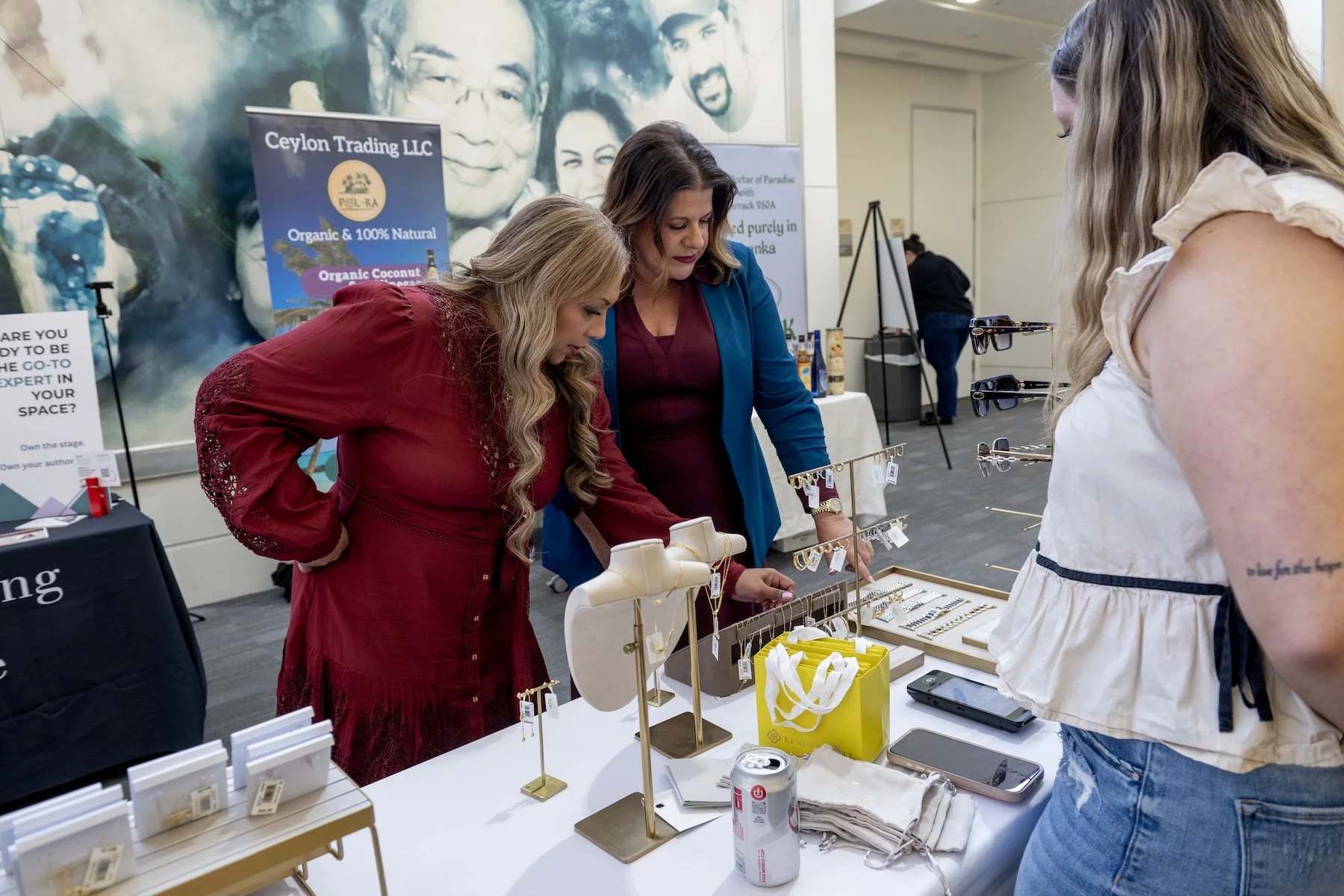 Three women shopping for jewelry at an indoor market stall. The stall displays various necklaces, rings, and earrings, with a woman in a white top looking at the jewelry and two women in red and blue dresses examining the items. There are smartphones, a soda can, and a yellow bag on the table.