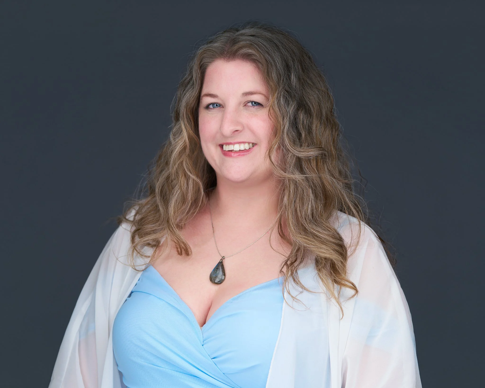 A woman with long, wavy brown hair, wearing a light blue top and a white sheer cover-up, smiling in front of a dark gray background.