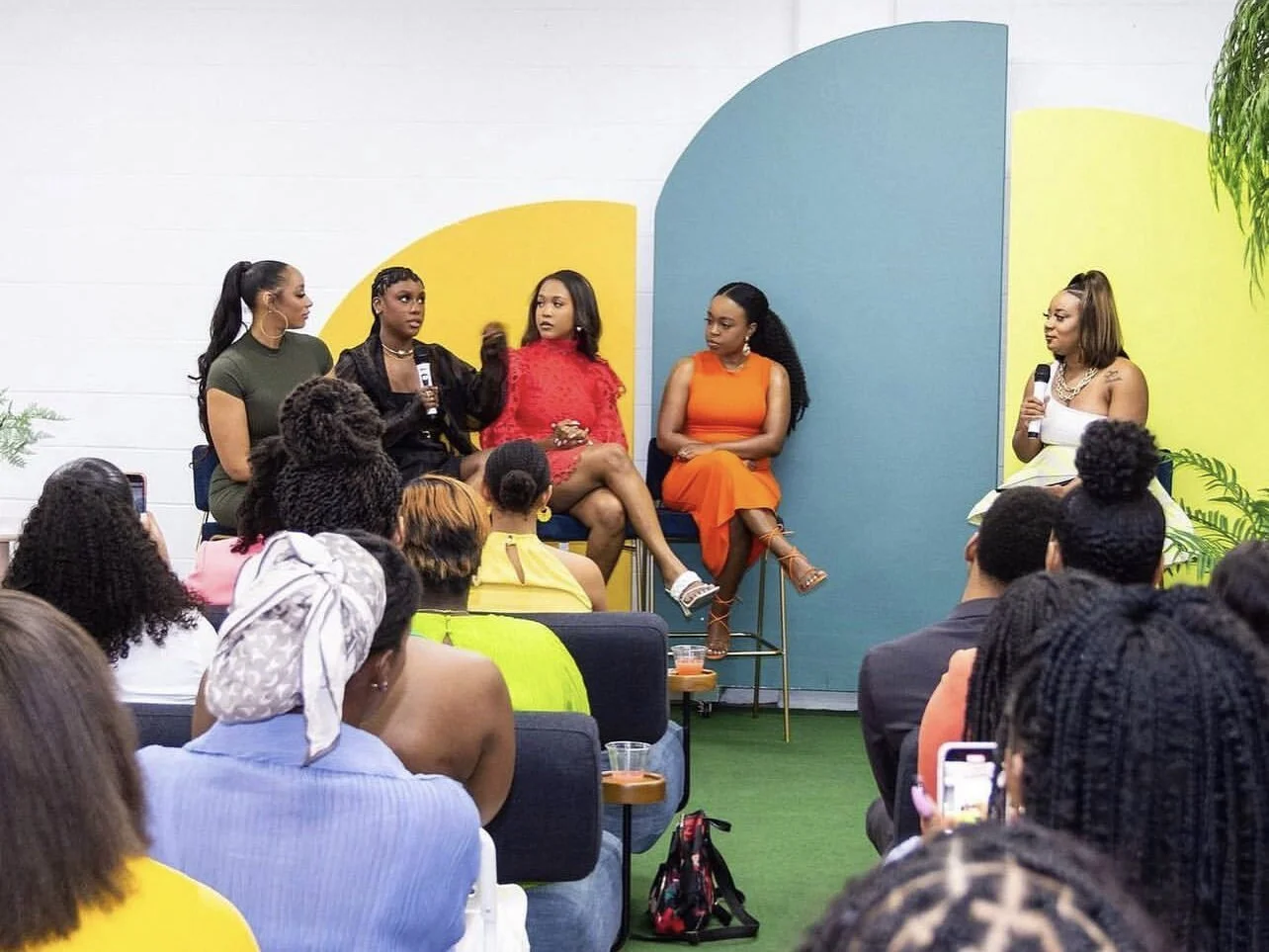 Women seated on stage participating in panel discussion, with a diverse audience watching, colorful backdrop with abstract shapes, and microphones in hand.