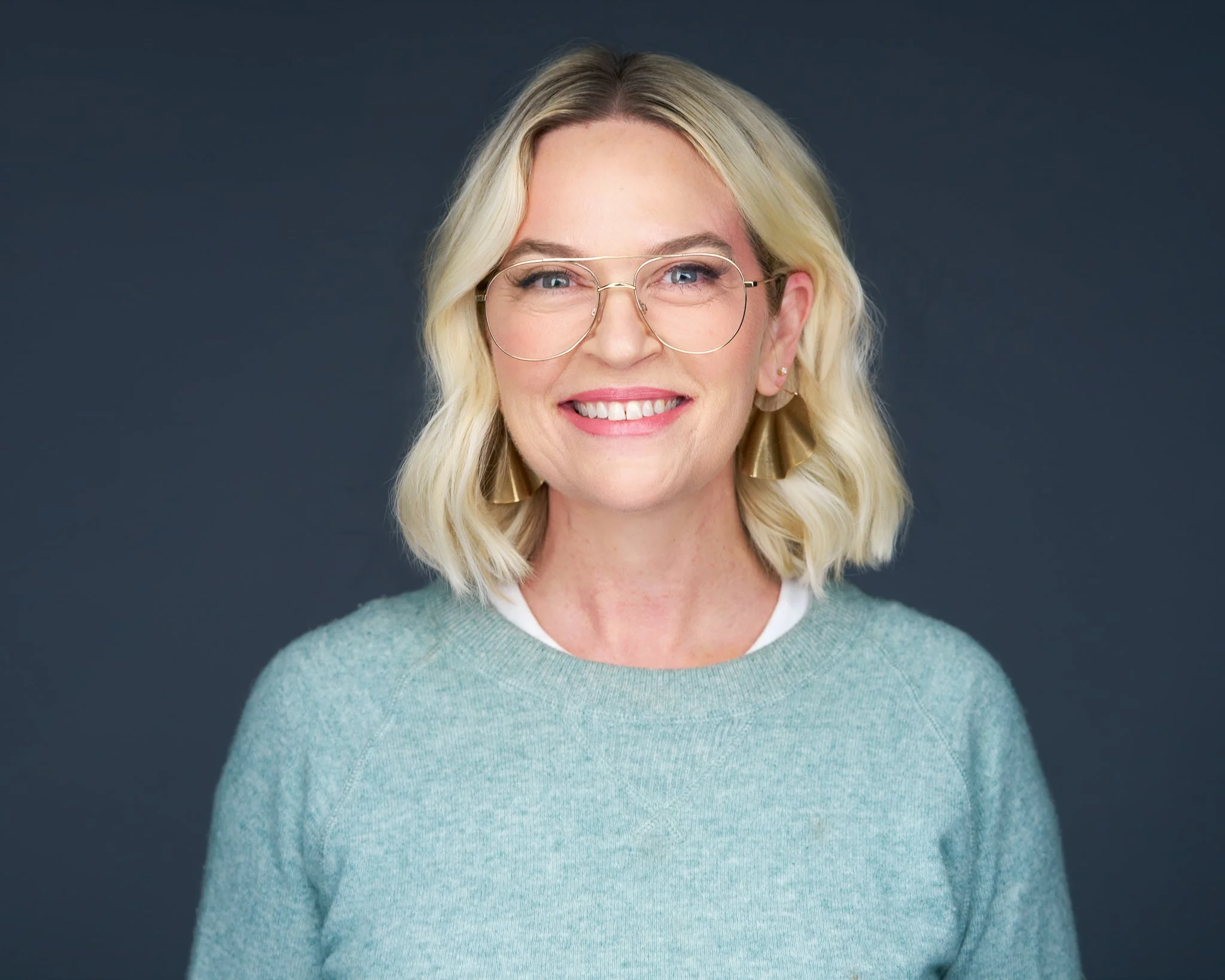 A smiling woman with blonde wavy hair, wearing round glasses, gold earrings, a light gray sweater, and a white shirt underneath, posed against a dark gray background.