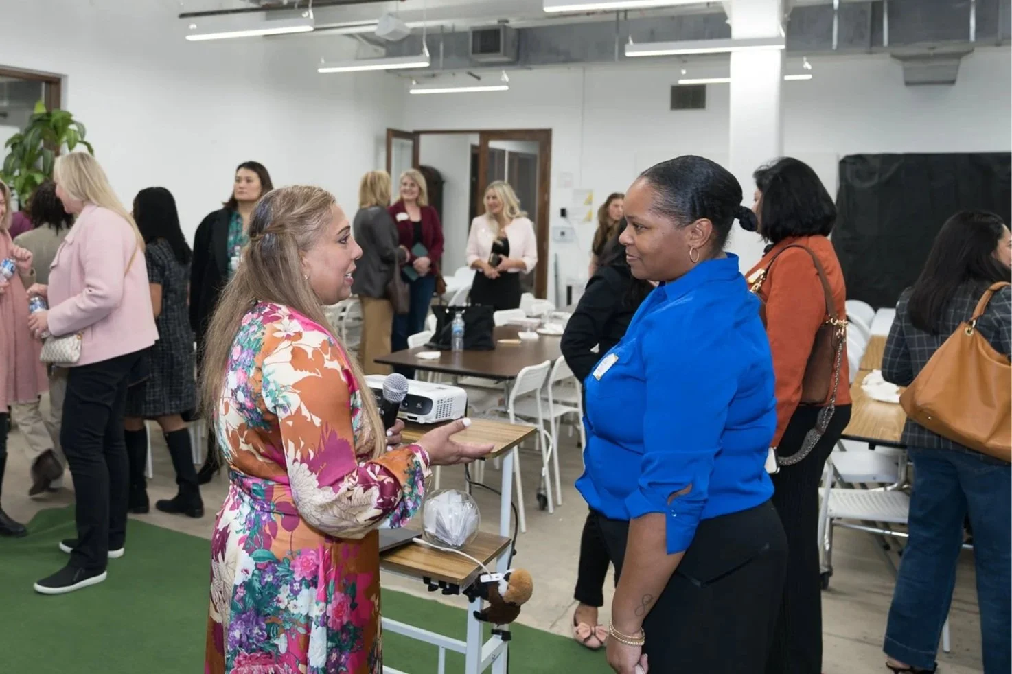 Two women conversing at an indoor networking event; one holding a microphone, the other wearing a blue blazer, with several people in the background.