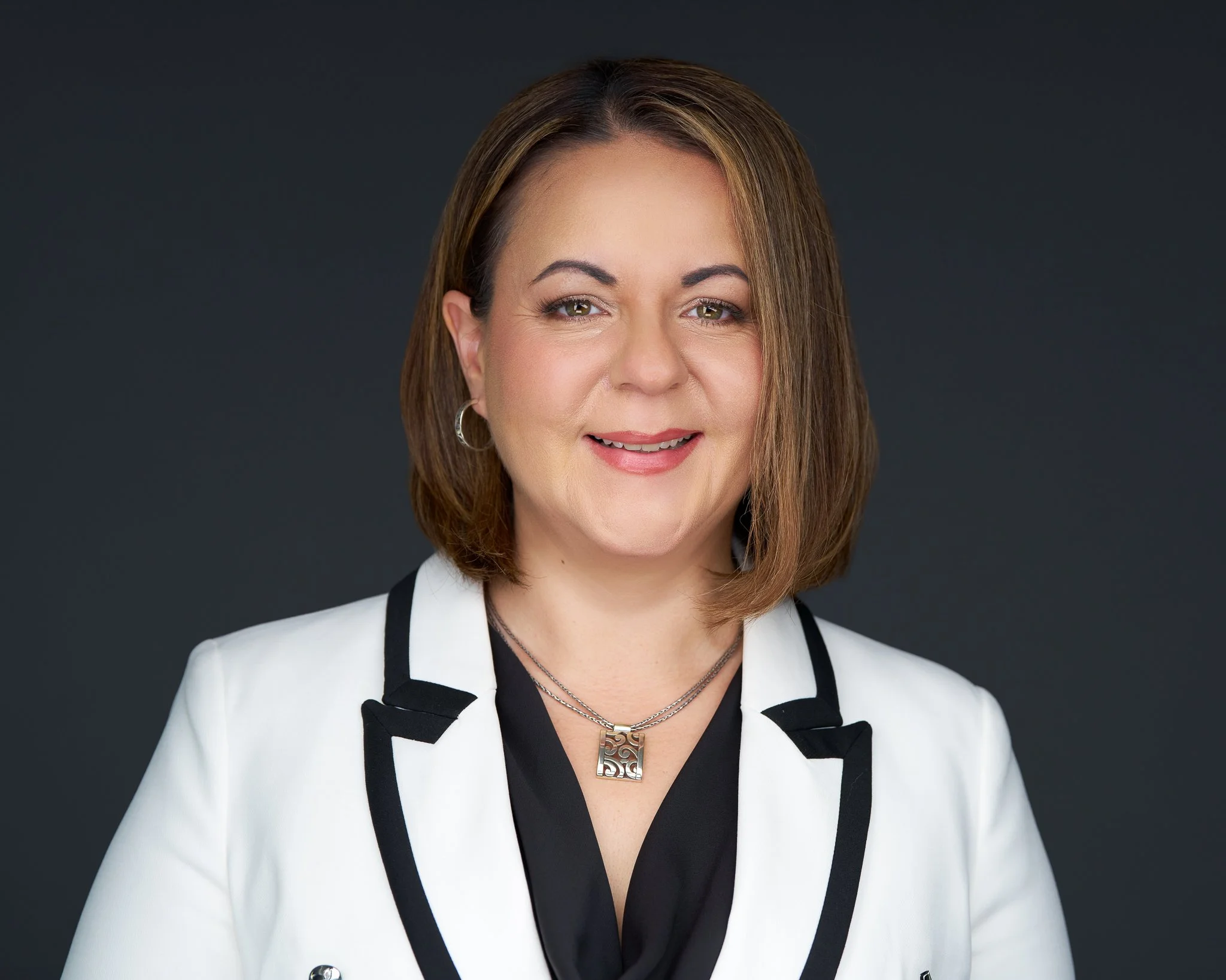 A woman with shoulder-length brown hair, wearing a white blazer with black accents, a black top, silver hoop earrings, and a silver necklace with a decorative pendant, smiling against a dark gray background.
