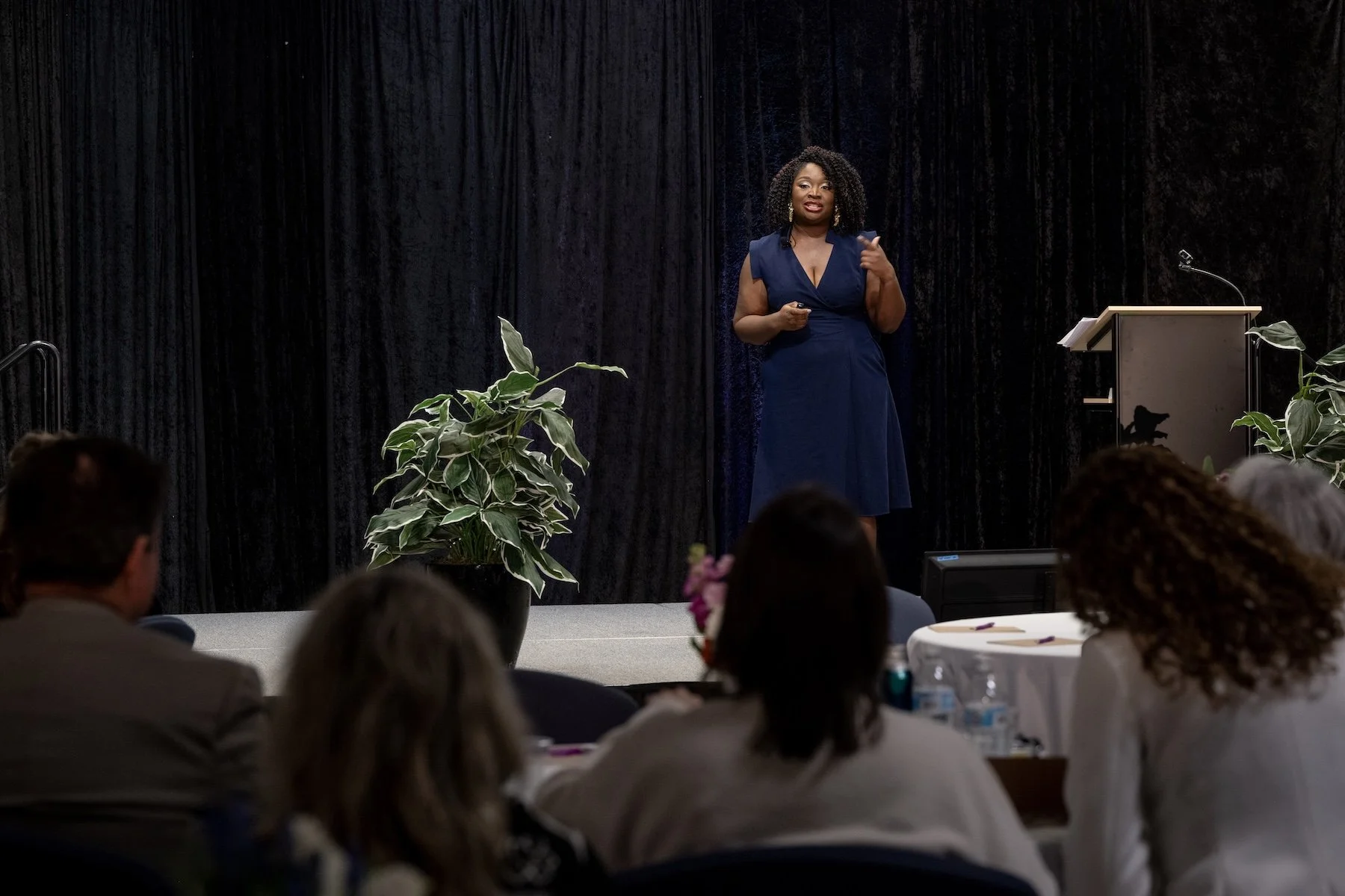 A woman in a blue dress giving a presentation on stage, with an audience listening.