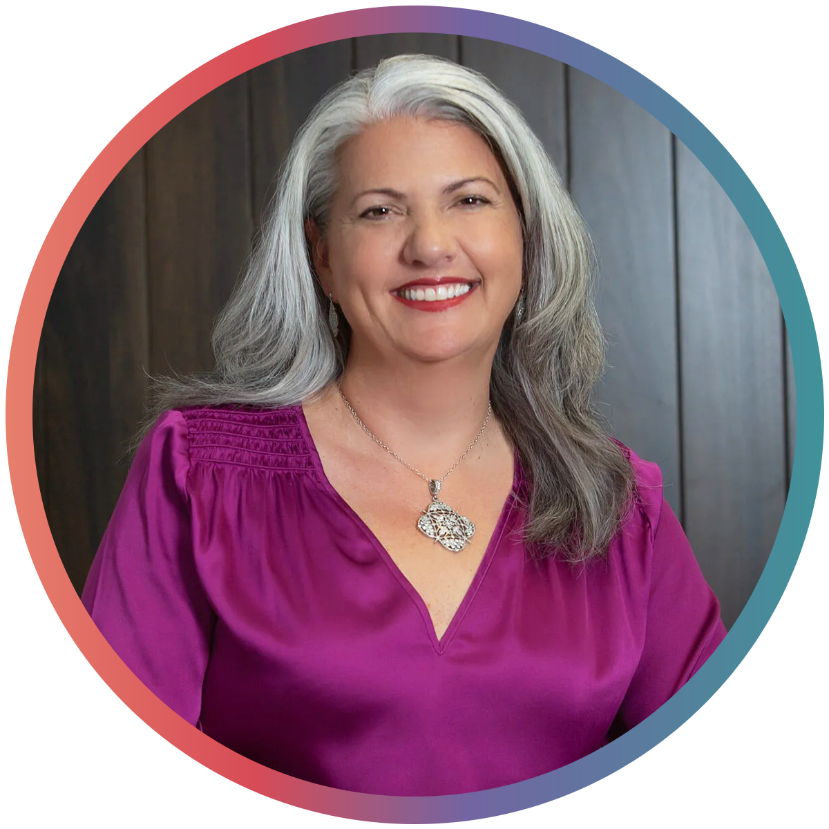 A smiling woman with long gray hair wearing a purple blouse and a silver necklace standing in front of a dark wooden wall.