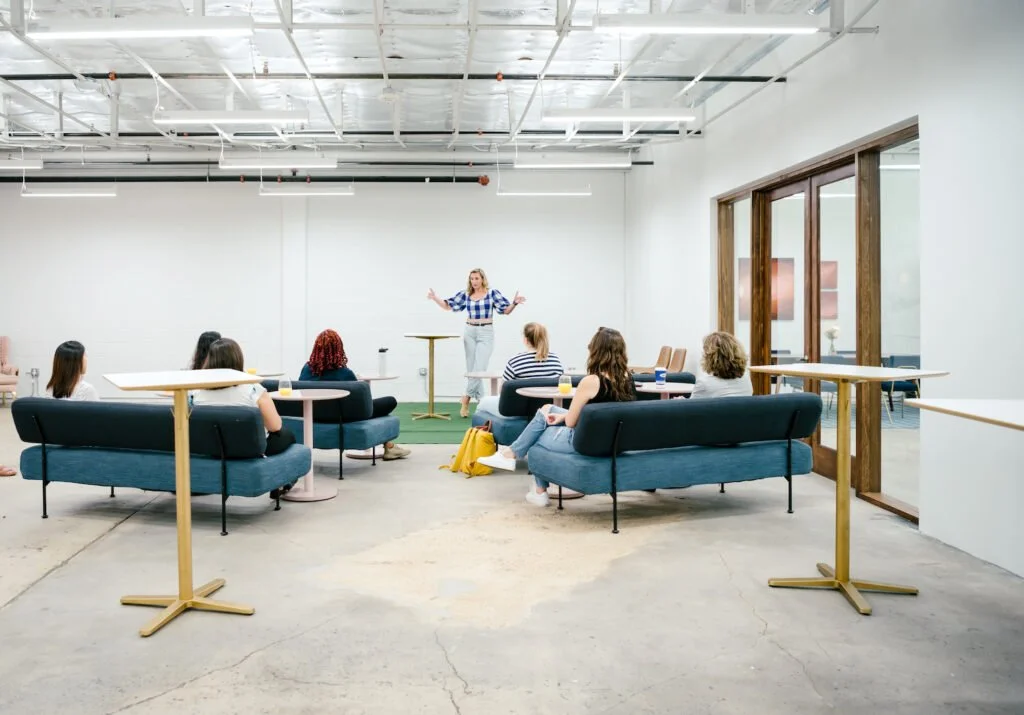 A woman giving a presentation to a group of eight women seated on sofas and chairs in a modern, minimalist conference room.
