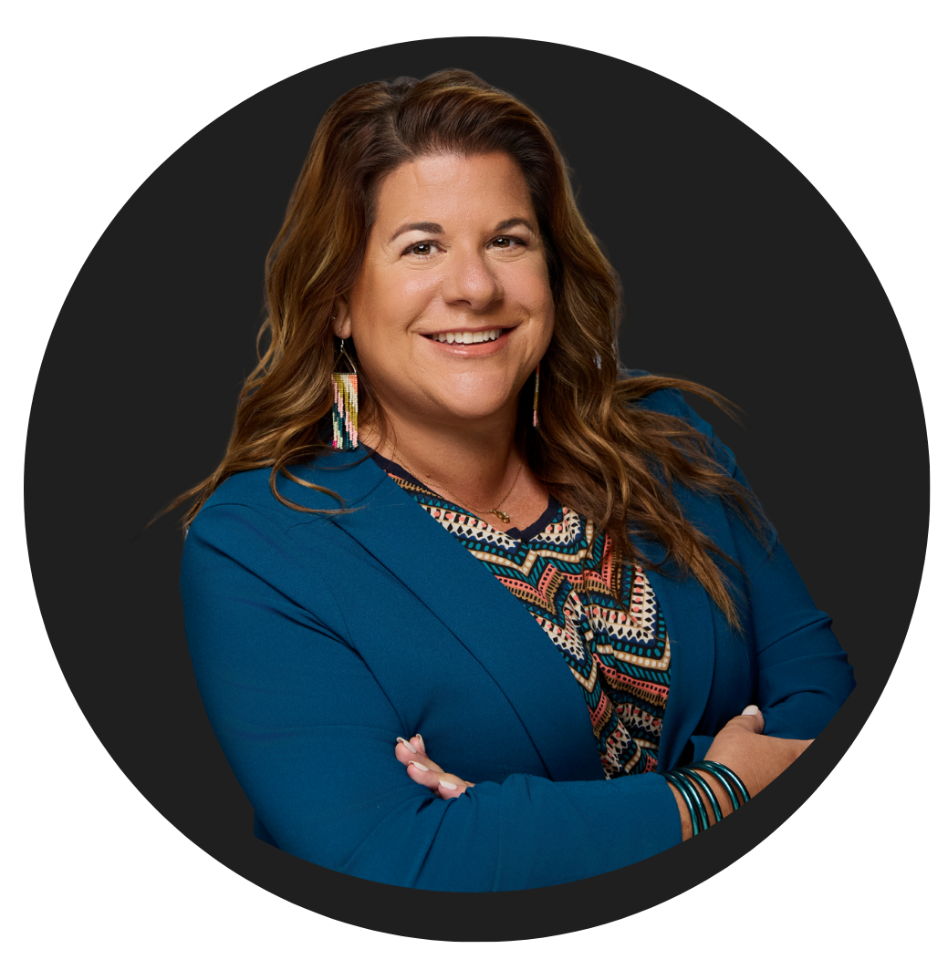 A woman smiling with crossed arms, wearing a blue blazer, colorful patterned top, and colorful earrings, standing against a black circular background.