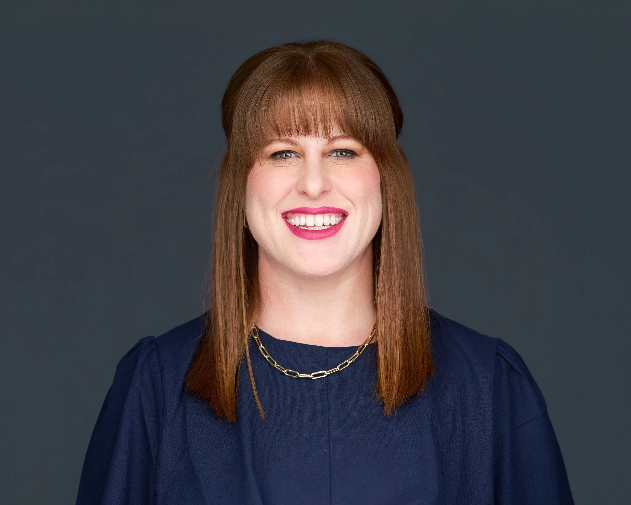 A woman with straight shoulder-length red hair, wearing a navy blue top and a gold chain necklace, smiling against a dark grey background.