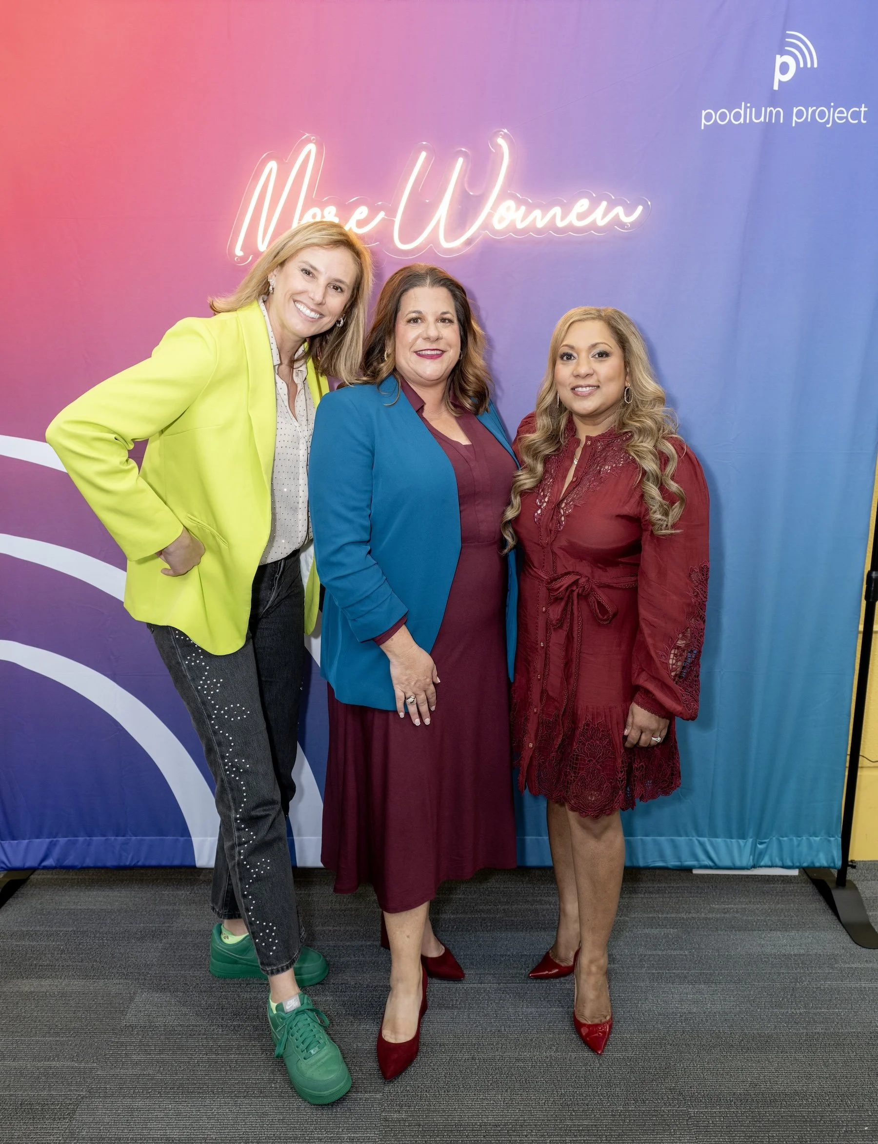 Three women, the Podium Project Founders and Sponsor, standing together in front of a colorful backdrop with the words 'More Women' in neon lights and 'podium project' logo in the corner.