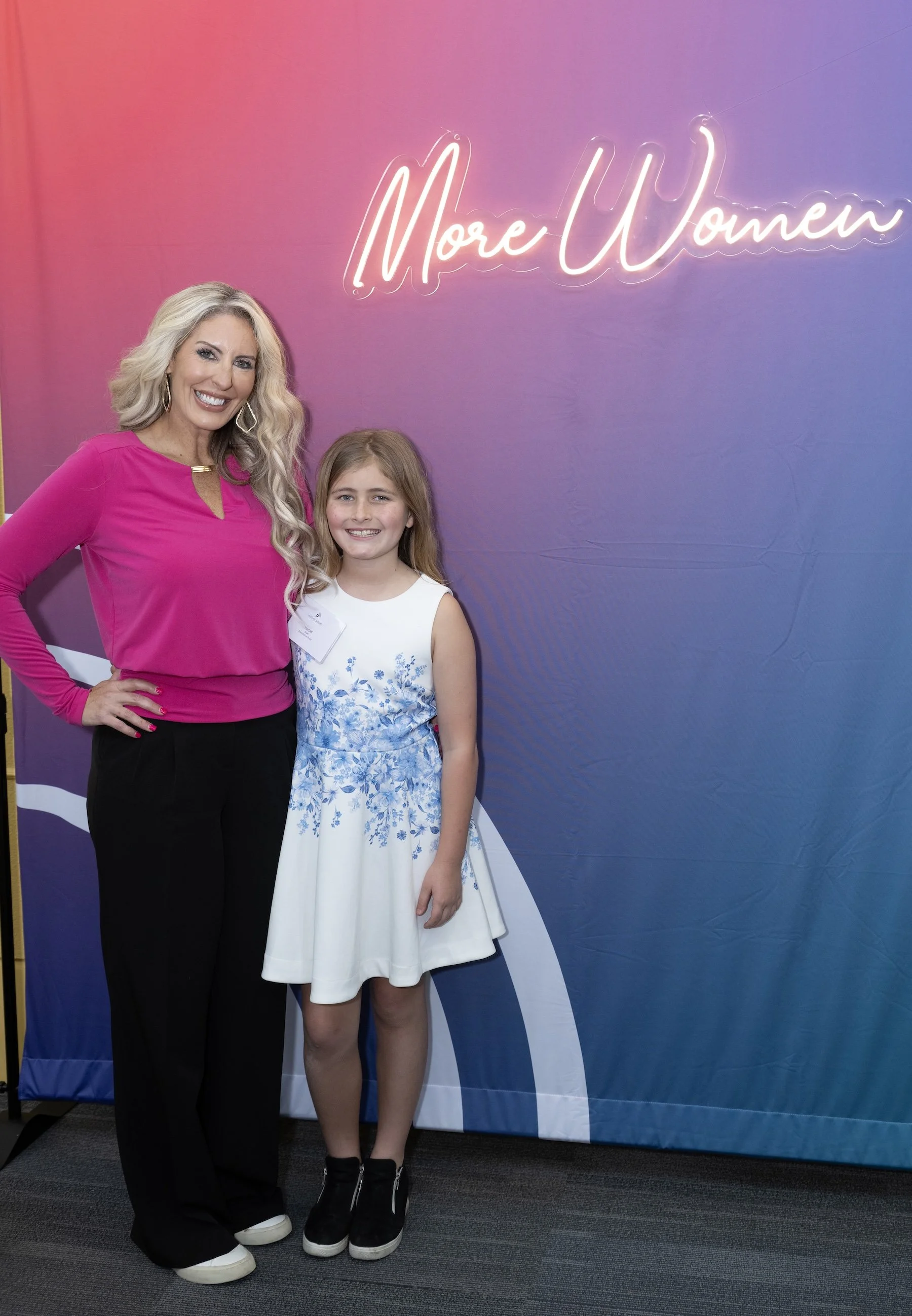 Woman and young girl smiling and posing in front of a neon sign that reads 'More Women'.