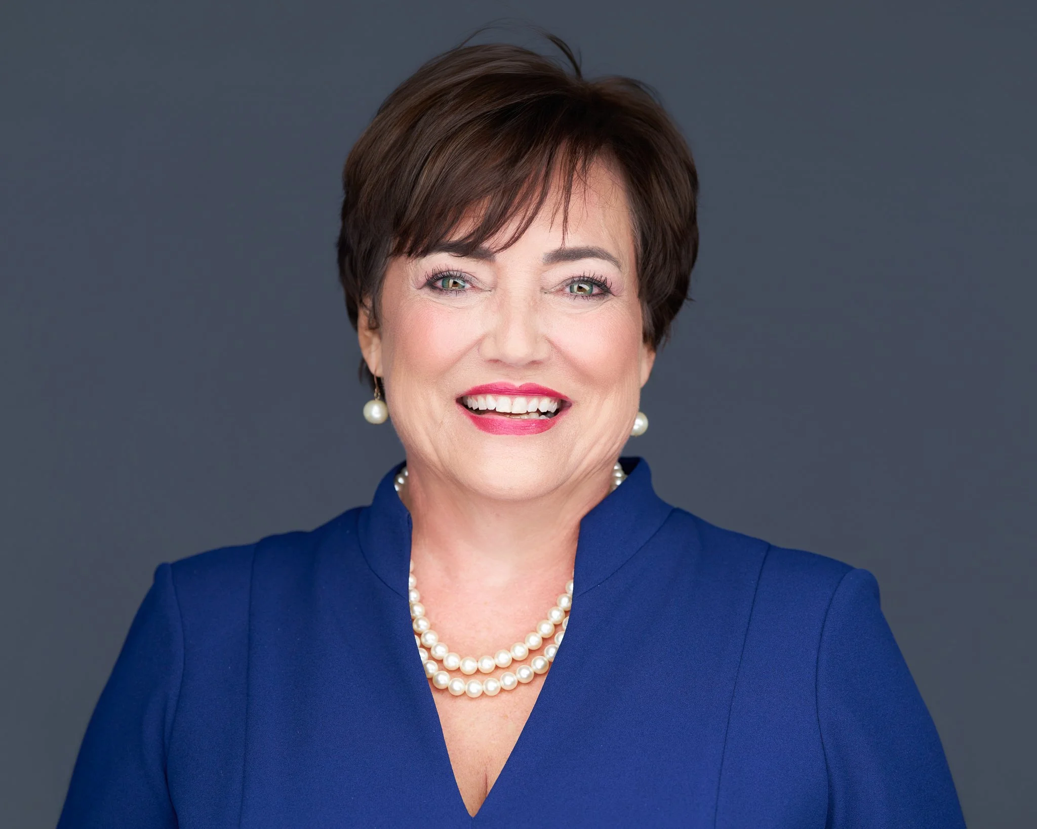 Portrait of a smiling woman in a blue blazer with pearl jewelry against a gray background.