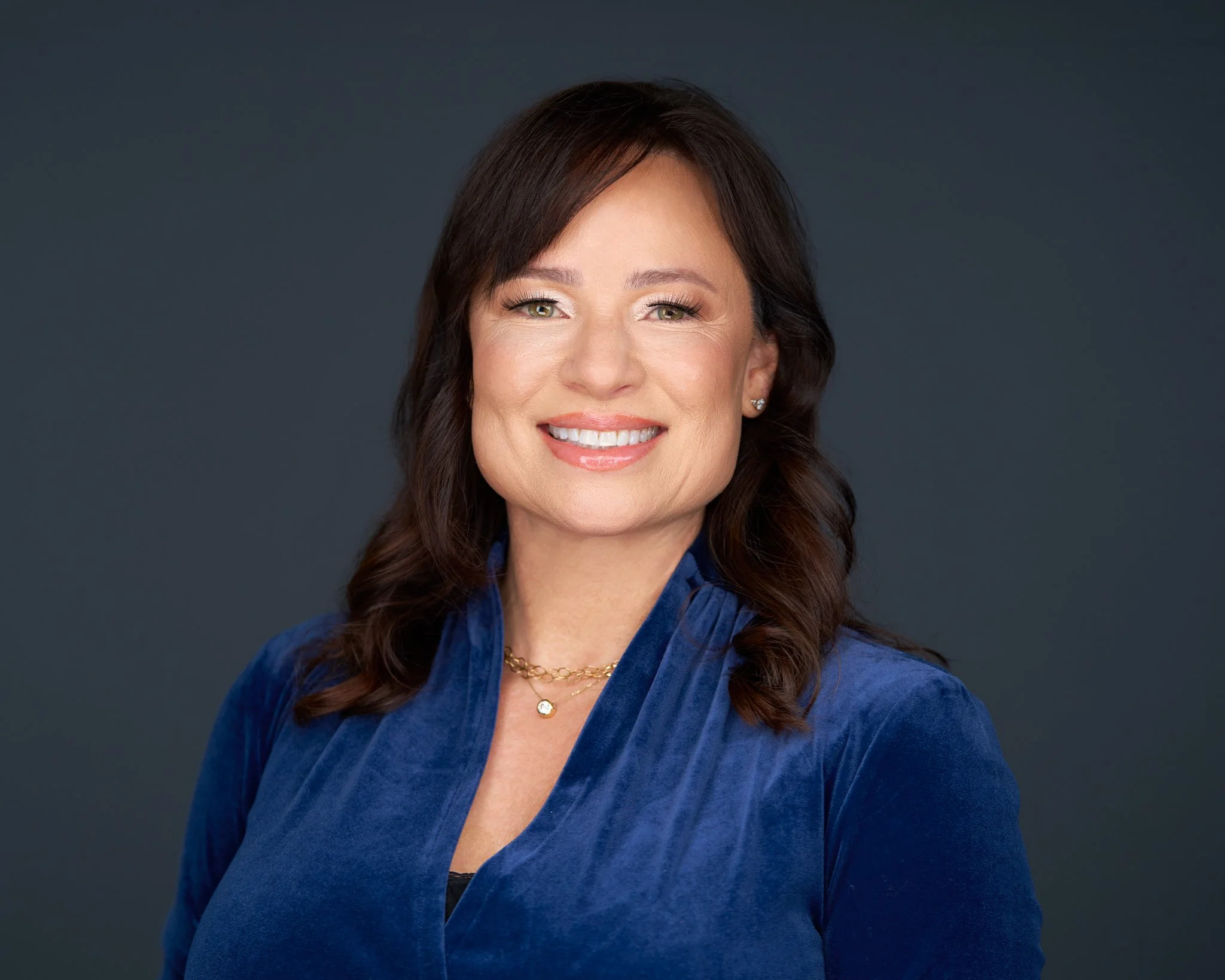 Headshot of a smiling woman with shoulder-length dark brown hair, wearing a blue velvet blazer and gold jewelry, against a dark gray background.