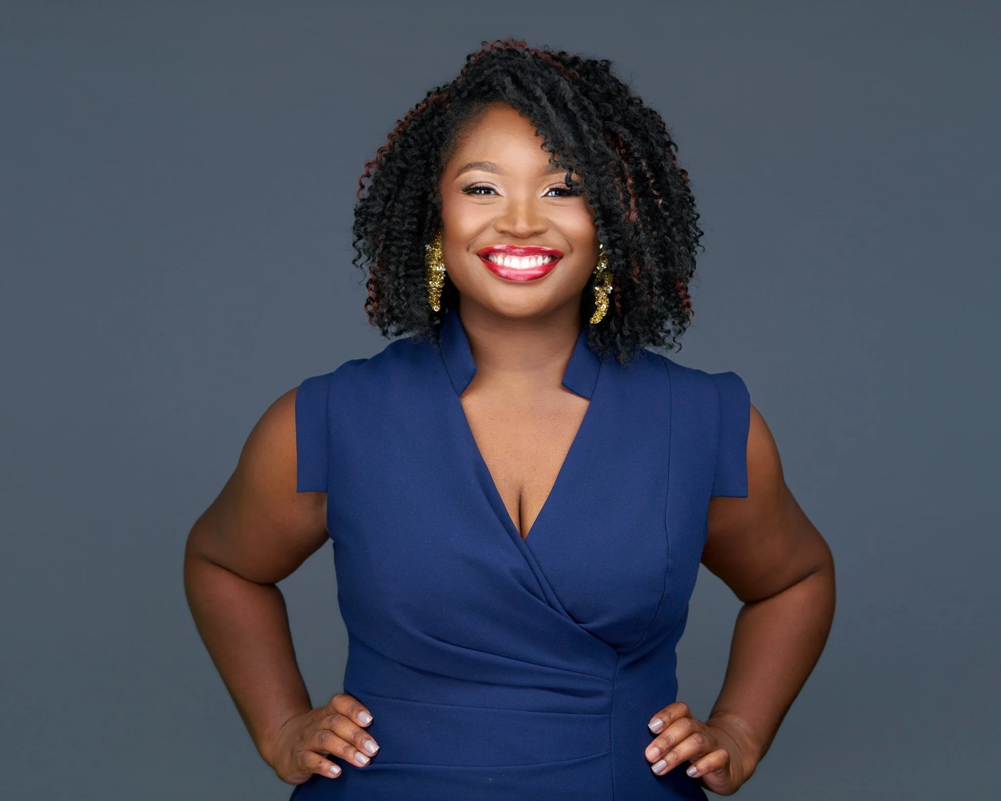 A woman with curly black hair wearing a blue sleeveless dress and gold earrings, smiling confidently with hands on hips against a gray background.