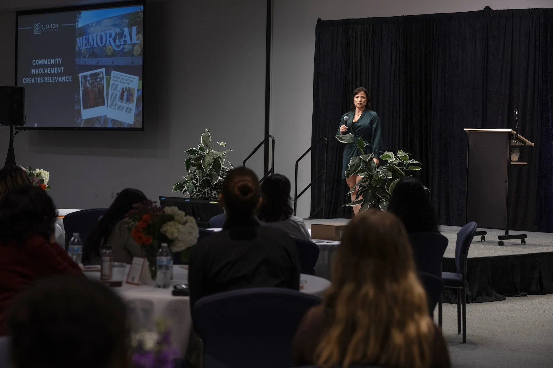 A woman in a dark green dress stands on a stage with a microphone, speaking to an audience in a conference room decorated with plants and flowers. A large screen to her left displays some text and images.