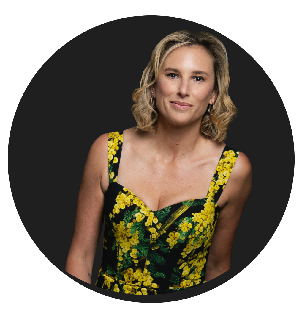 A woman with blonde hair styled in loose waves, wearing a black dress with a yellow floral pattern, standing against a dark background.