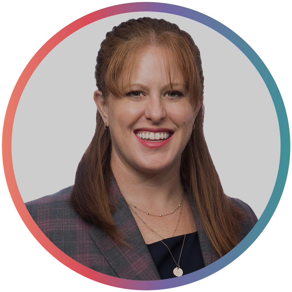 A professional woman with shoulder-length red hair smiling, wearing a dark blazer with a plaid pattern and layered necklaces, against a light grey background.