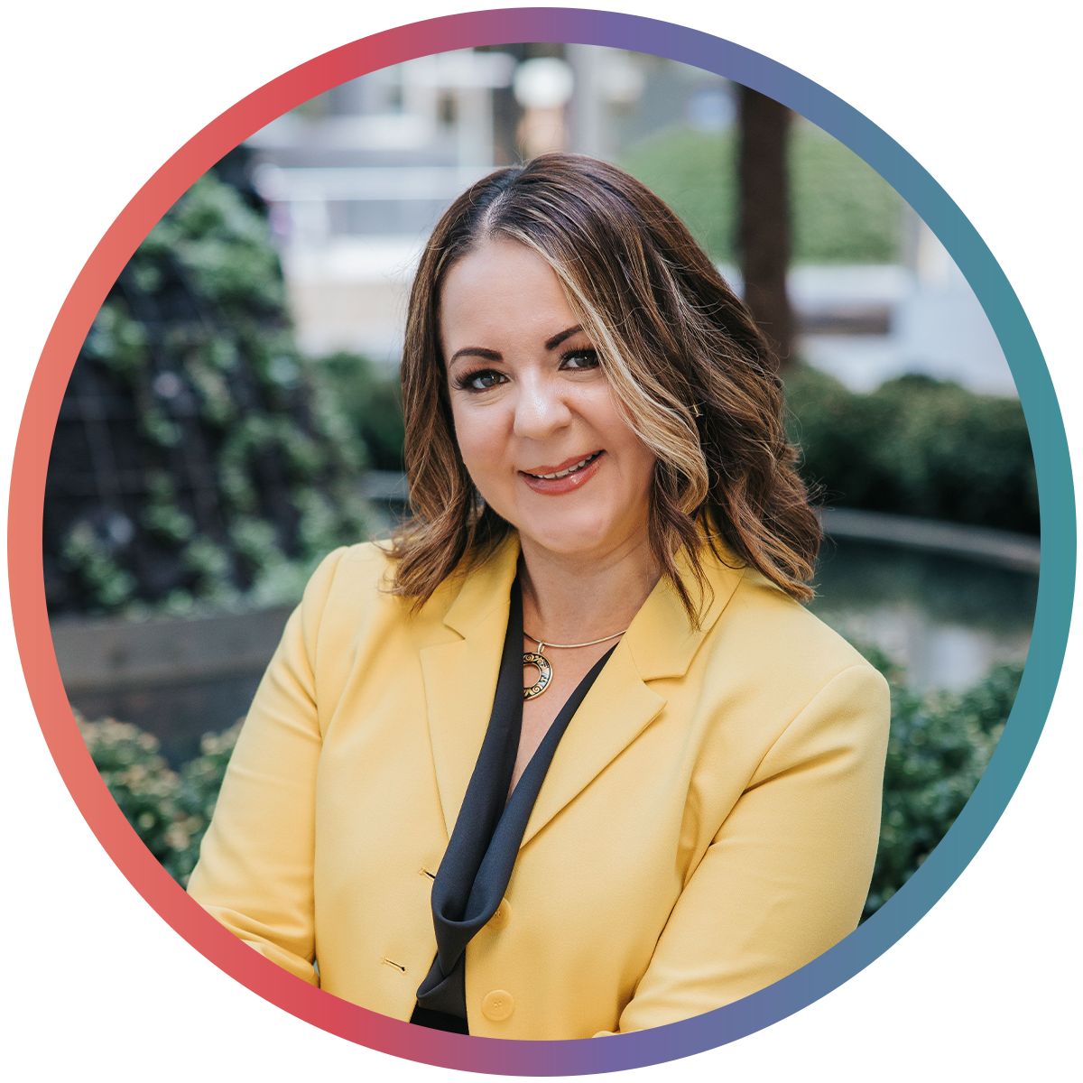 A woman with shoulder-length brown hair and a bright smile, wearing a yellow blazer and a necklace, standing outdoors with greenery in the background.